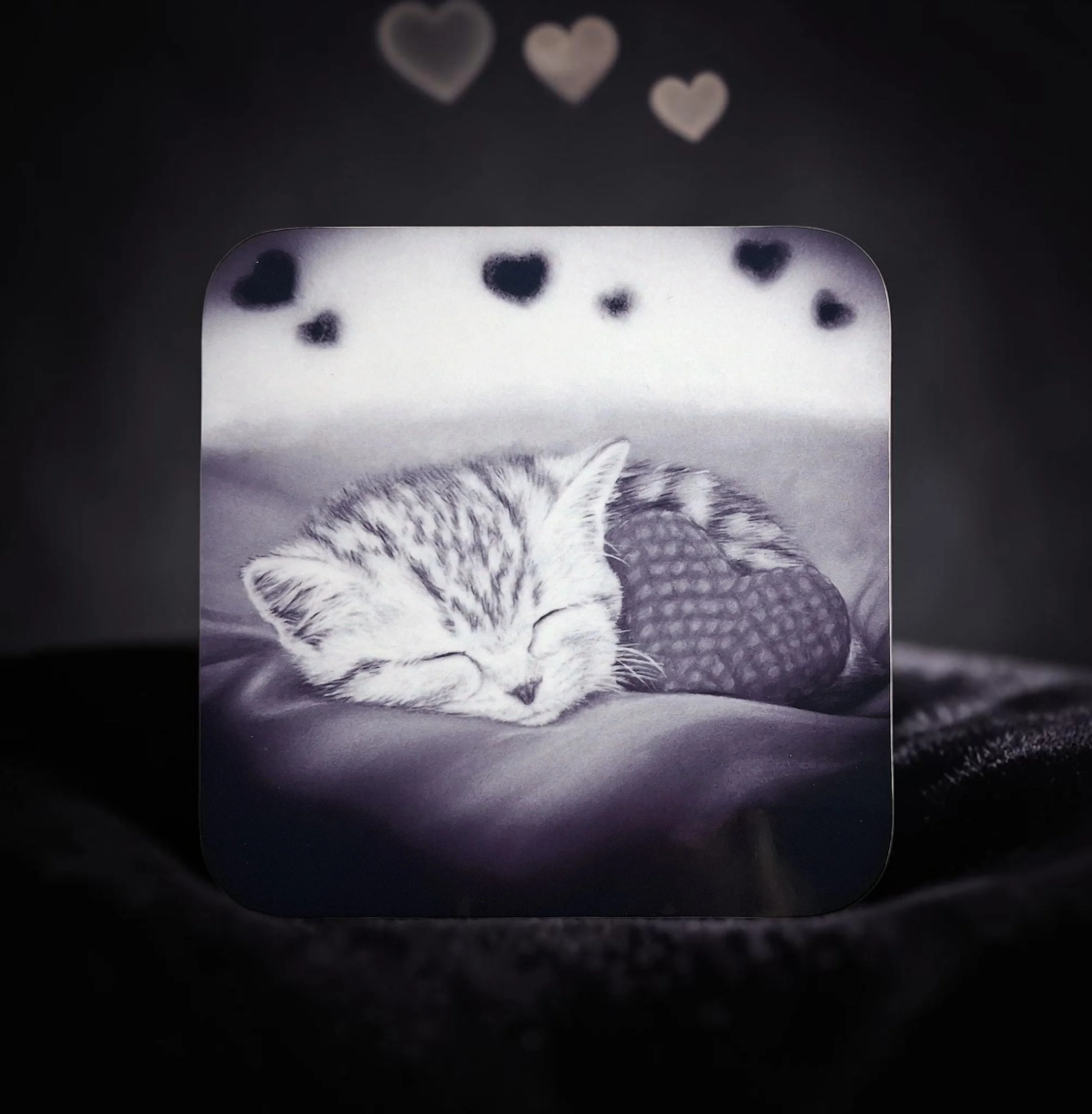 Hand drawn artwork printed onto hardboard coaster, in black and white glossy finish of a sleeping kitten snuggled up with a heart cushion