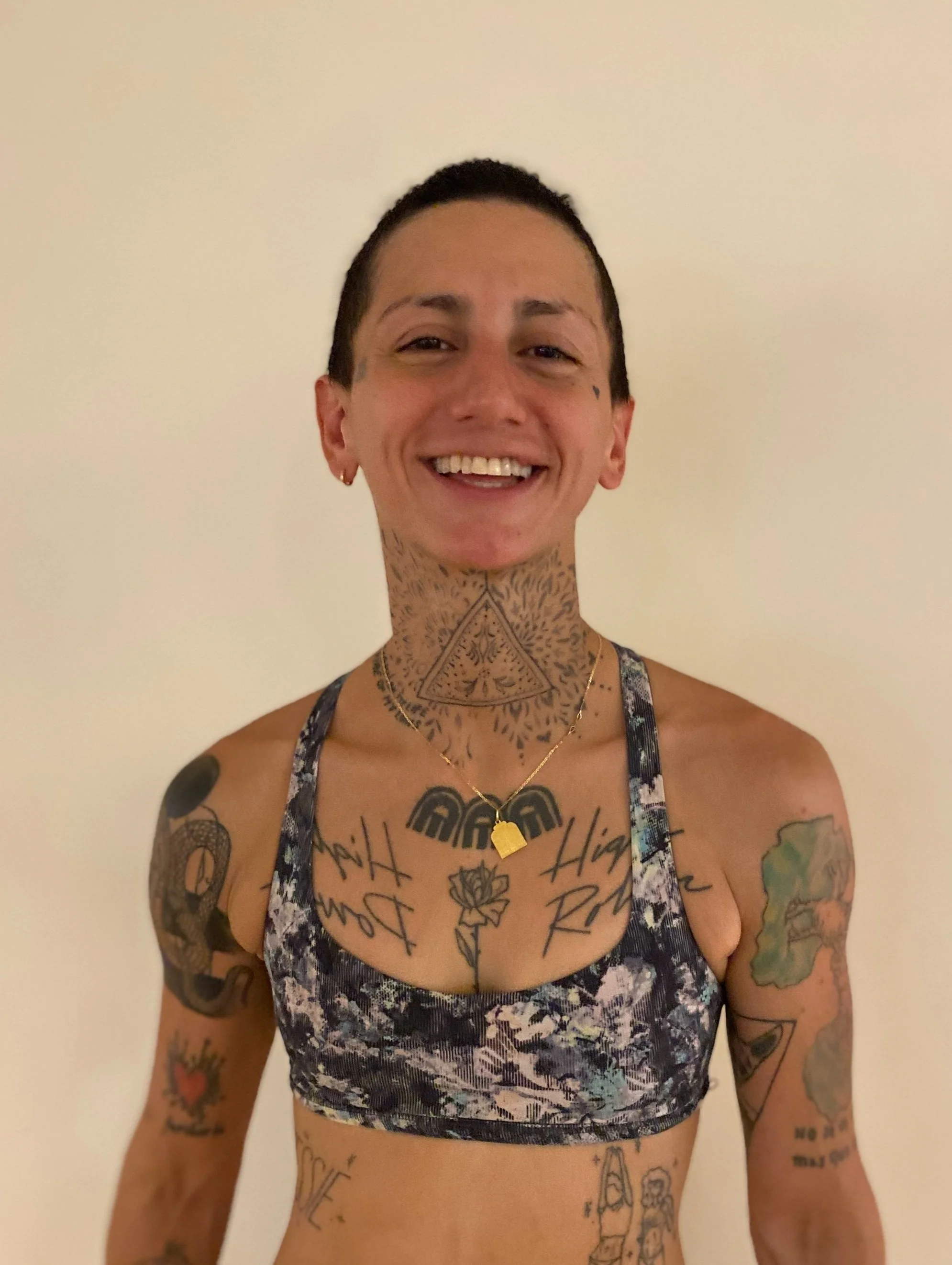 A person with short dark hair smiling, wearing a floral sports bra, with numerous tattoos on their arms, chest, and neck, and a gold necklace with a pendant.