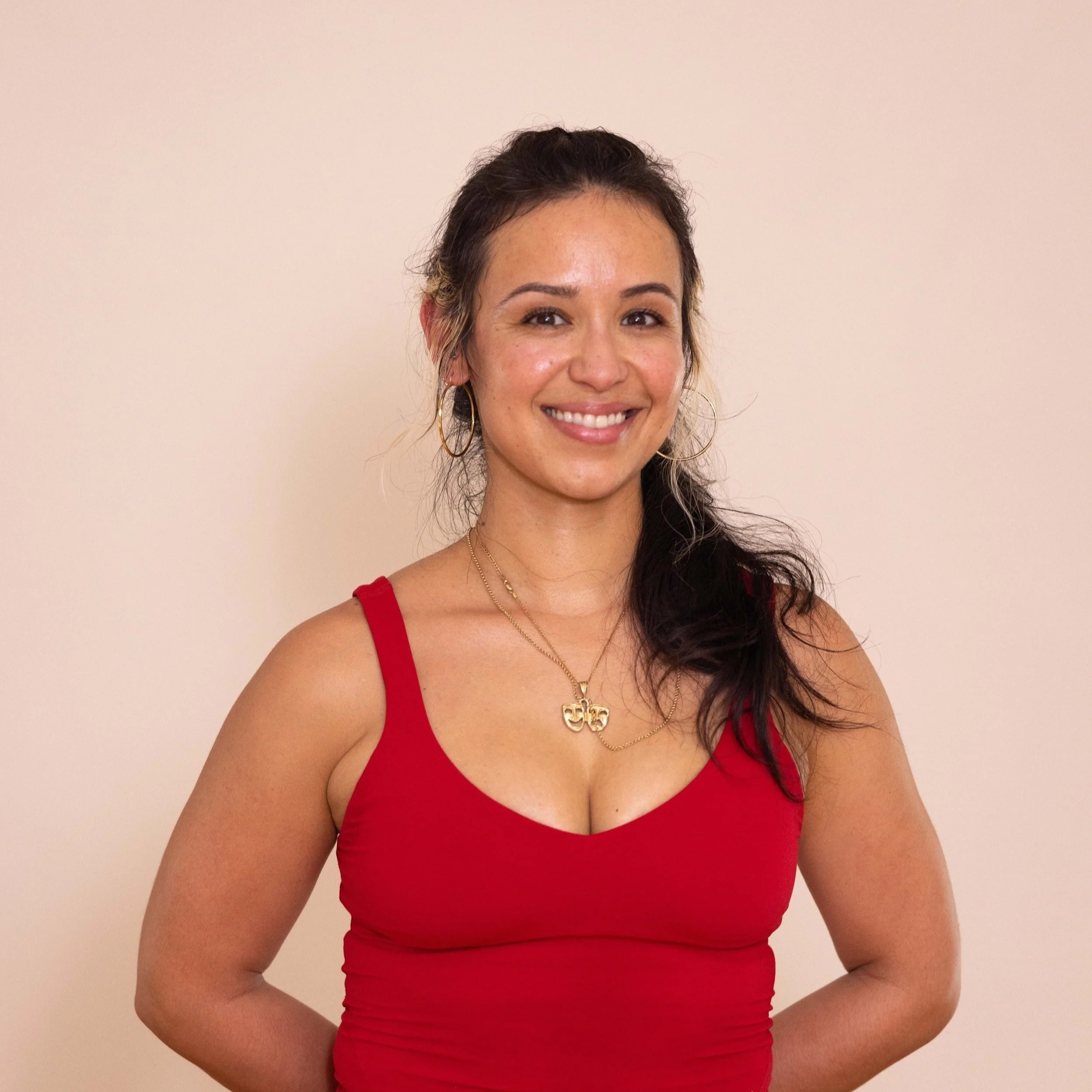 A smiling woman wearing a red tank top, gold earrings, and gold necklaces standing against a plain beige wall.