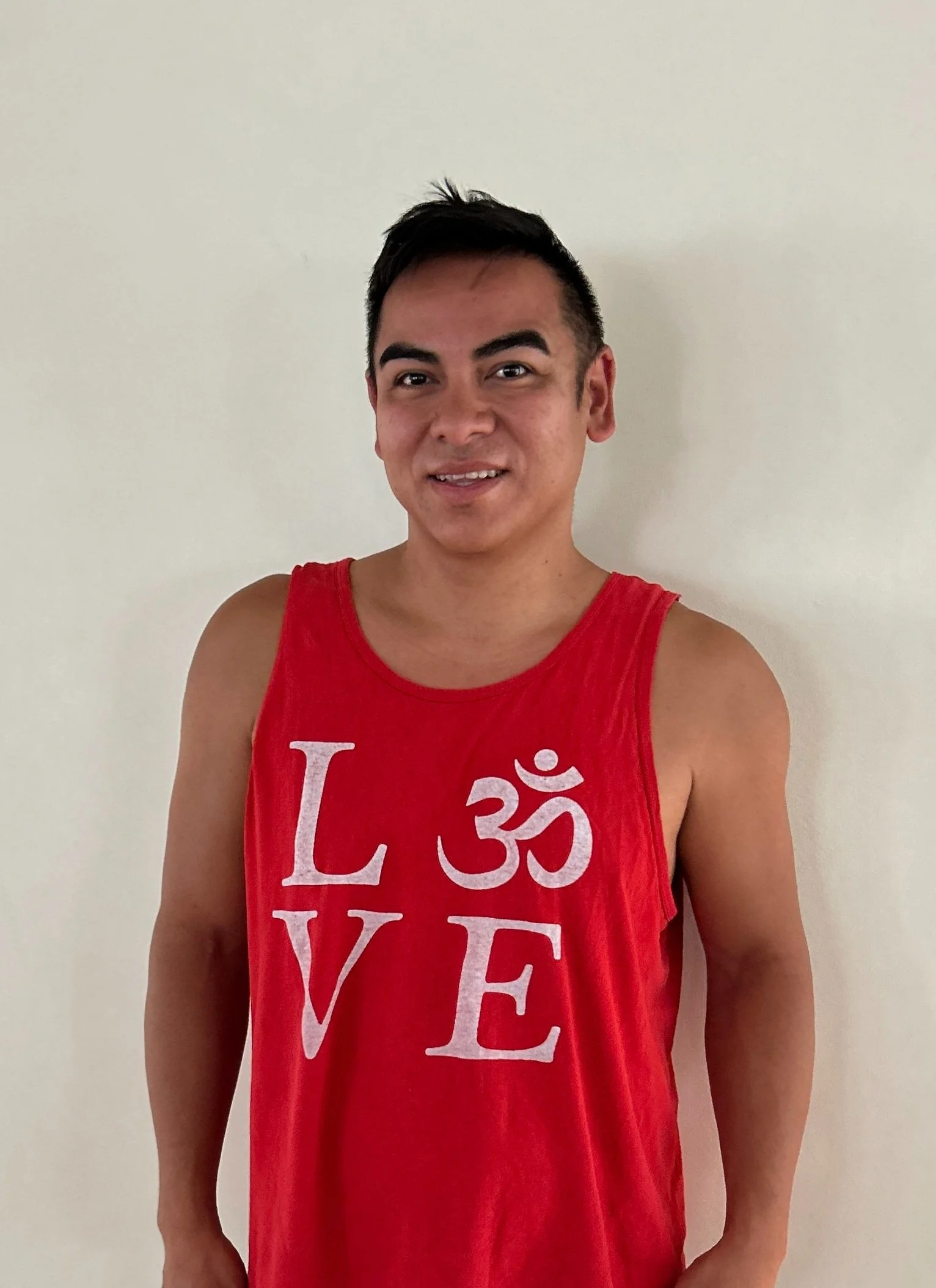 A young man with short dark hair and light brown skin wearing a red sleeveless shirt with the word love and an Om symbol printed on it, standing against a plain beige wall.