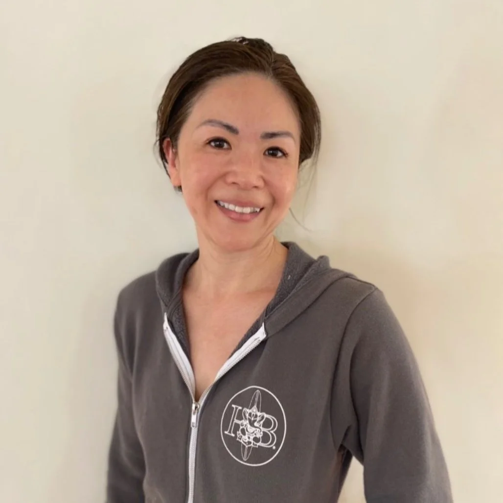 Smiling woman with short brown hair wearing a gray zip-up hoodie with a logo on the left chest, standing against a plain light-colored wall.