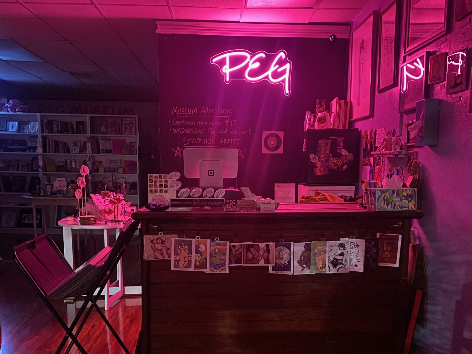 A museum reception area illuminated with pink neon lights that spell 'PEG.' There are photos and artwork displayed on the wall, a chalkboard with admission prices, and shelves filled with books and art supplies. Small tables with decorative items and chairs are also present.