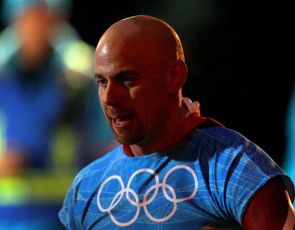A male athlete in a blue shirt with Olympic rings, looking down, with a blurred background.