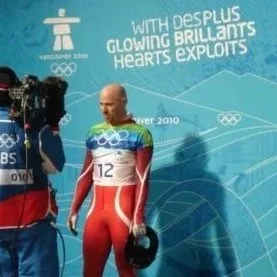 Olympic athlete in a red and blue uniform holding a helmet, standing in front of a Vancouver 2010 Olympic backdrop. A camera crew films him.