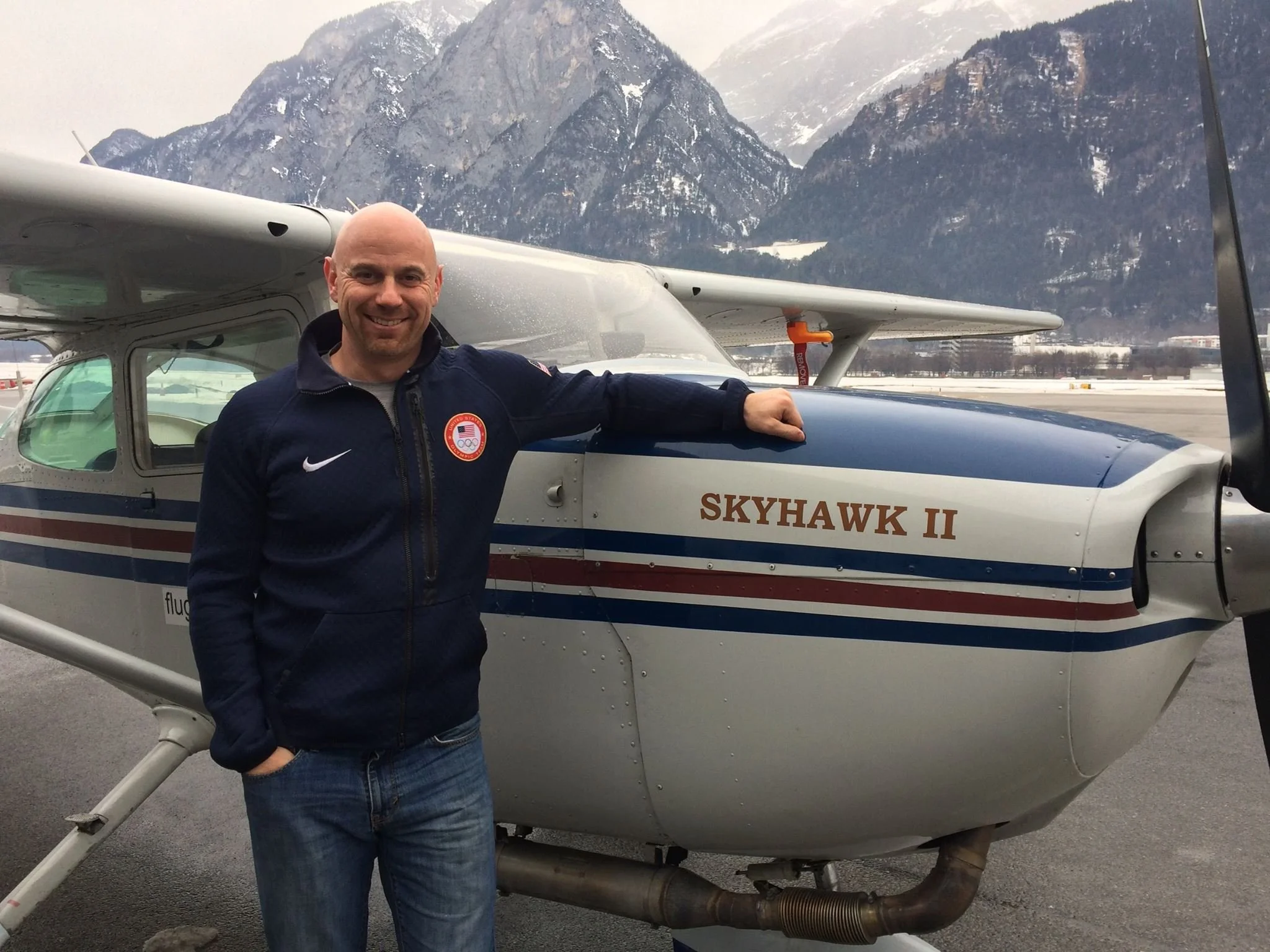 A man in a navy jacket with an Olympic emblem on the chest is standing next to a small aircraft named Skyhawk II, with snow-covered mountains in the background.