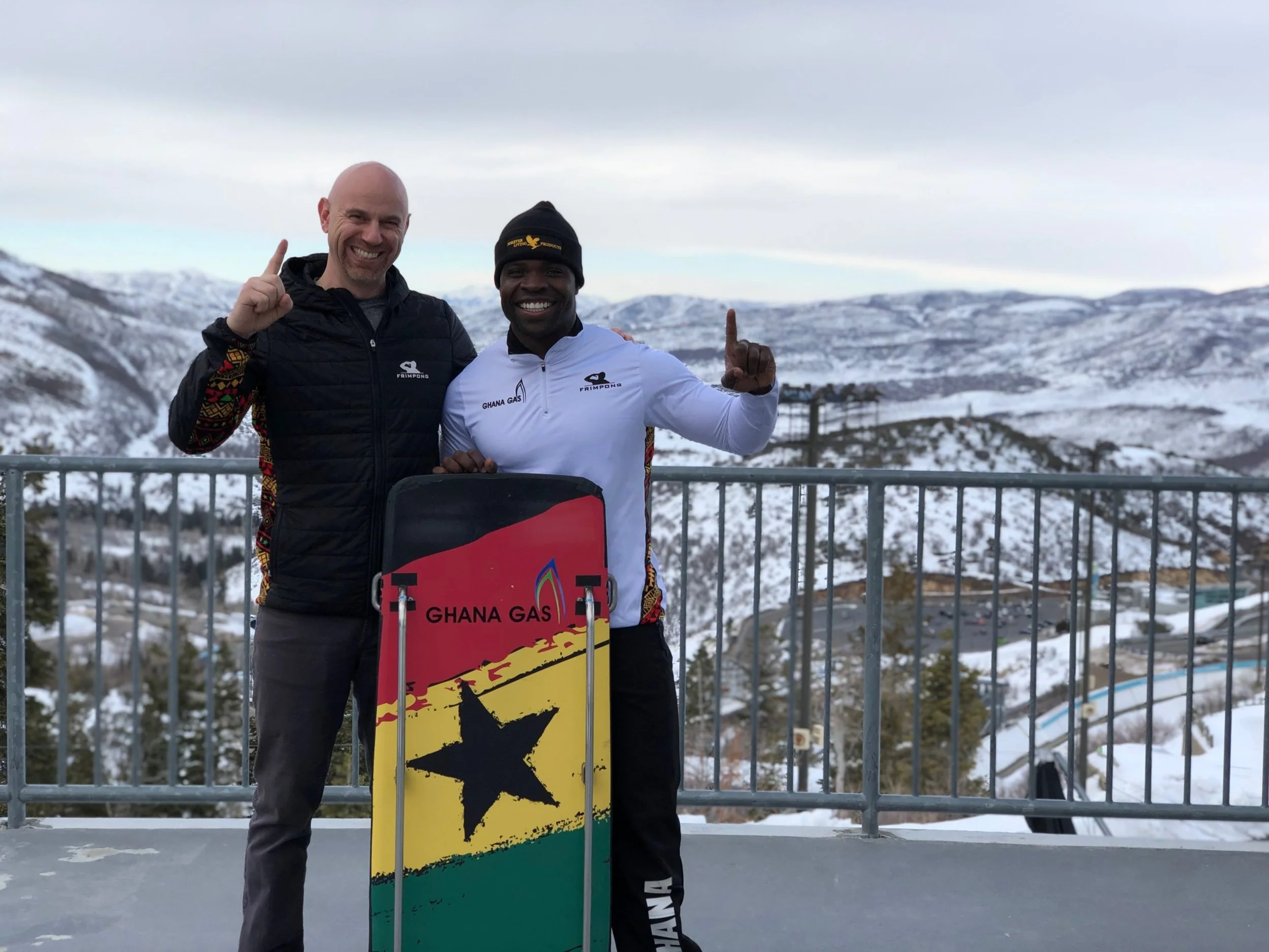 Two men standing outdoors in snowy mountains, smiling and holding a skateboard with the Ghana Gas logo, dressed in athletic clothing.