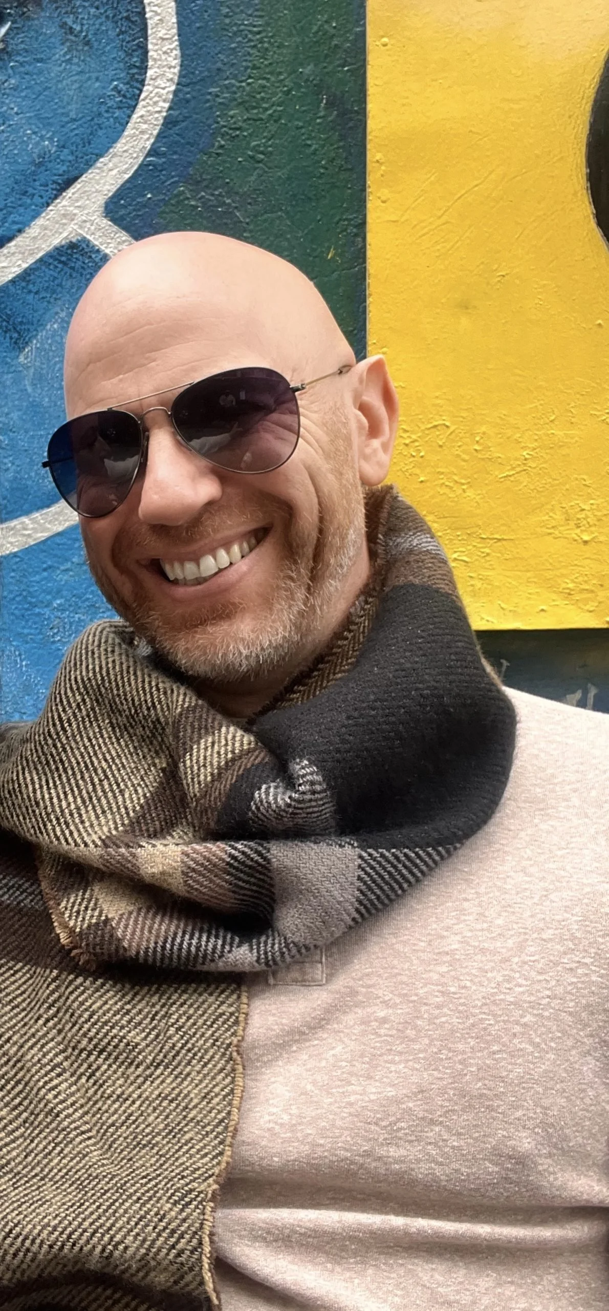A smiling man with a bald head, wearing dark sunglasses, a brown and beige checkered scarf, and a light-colored sweater. He is standing in front of a colorful, abstract painted wall with yellow, green, black, blue, and white sections.