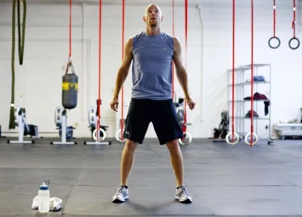 A man in athletic wear standing in a gym, surrounded by exercise equipment including rowing machines, punching bags, and gymnastic rings.