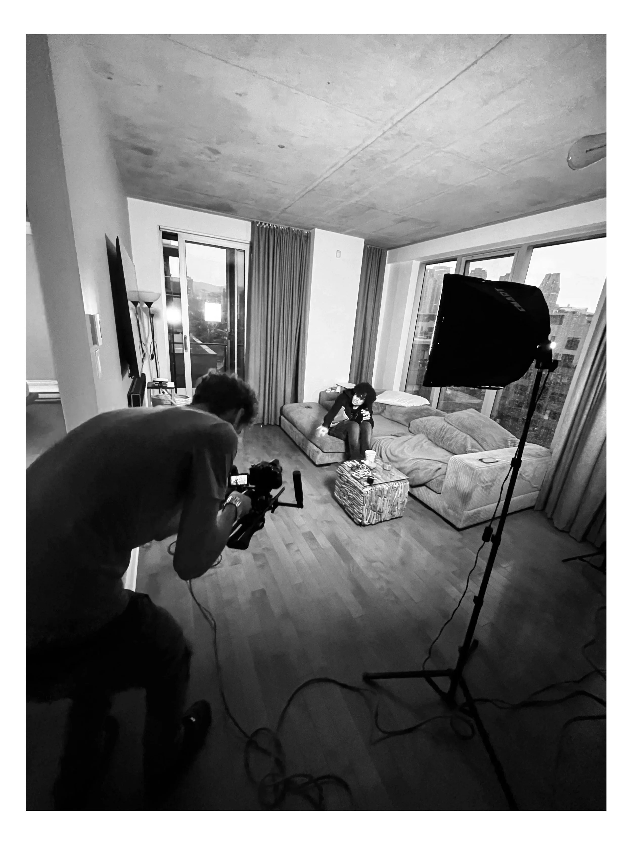 Une scène de tournage dans un salon moderne avec deux personnes, une cabine d'éclairage et un équipement de caméra, deux grandes fenêtres laissent entrer la lumière naturelle.