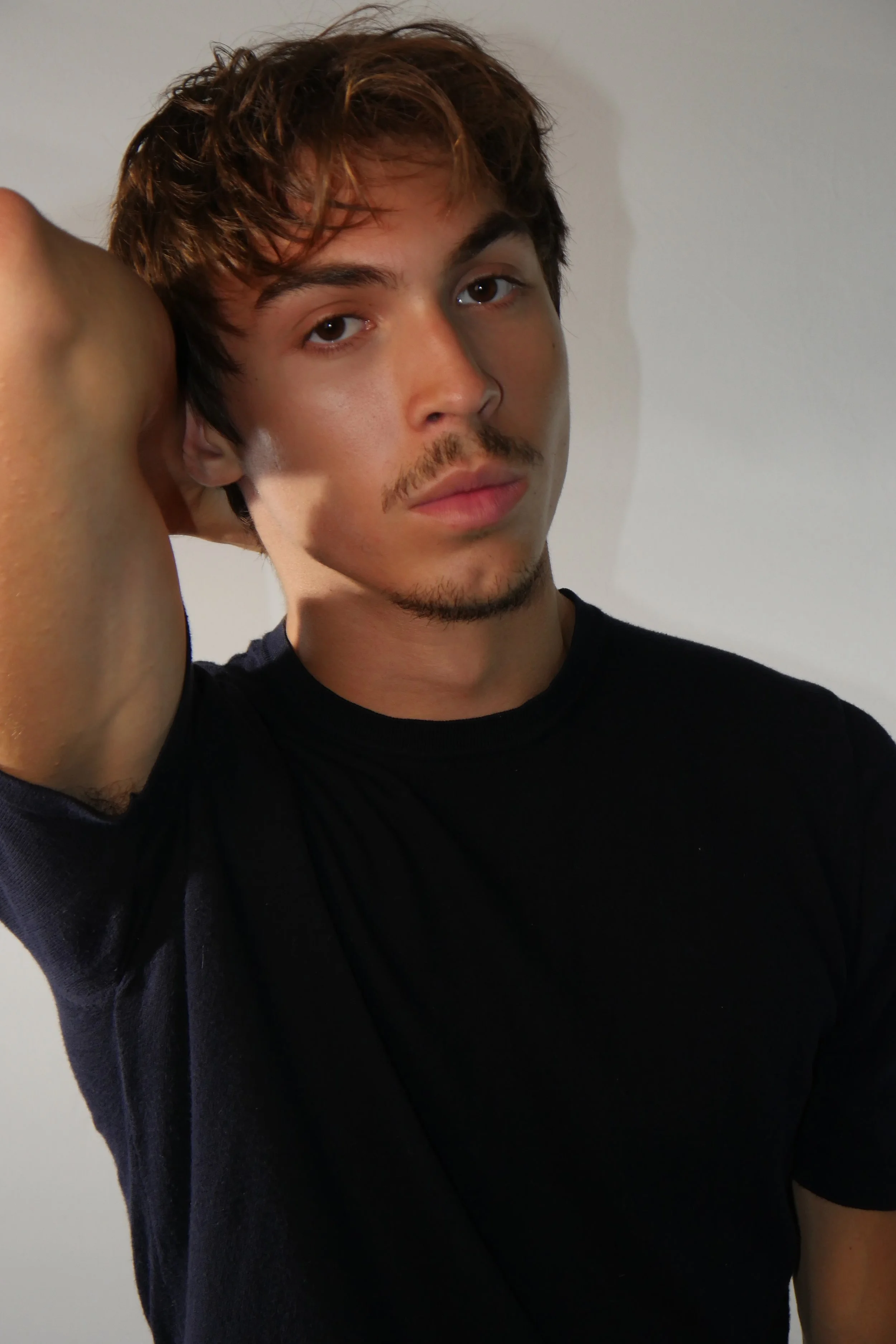 Portrait d'un jeune homme aux cheveux bruns, portant un t-shirt noir, avec un bras levé derrière la tête, posant devant un mur blanc.