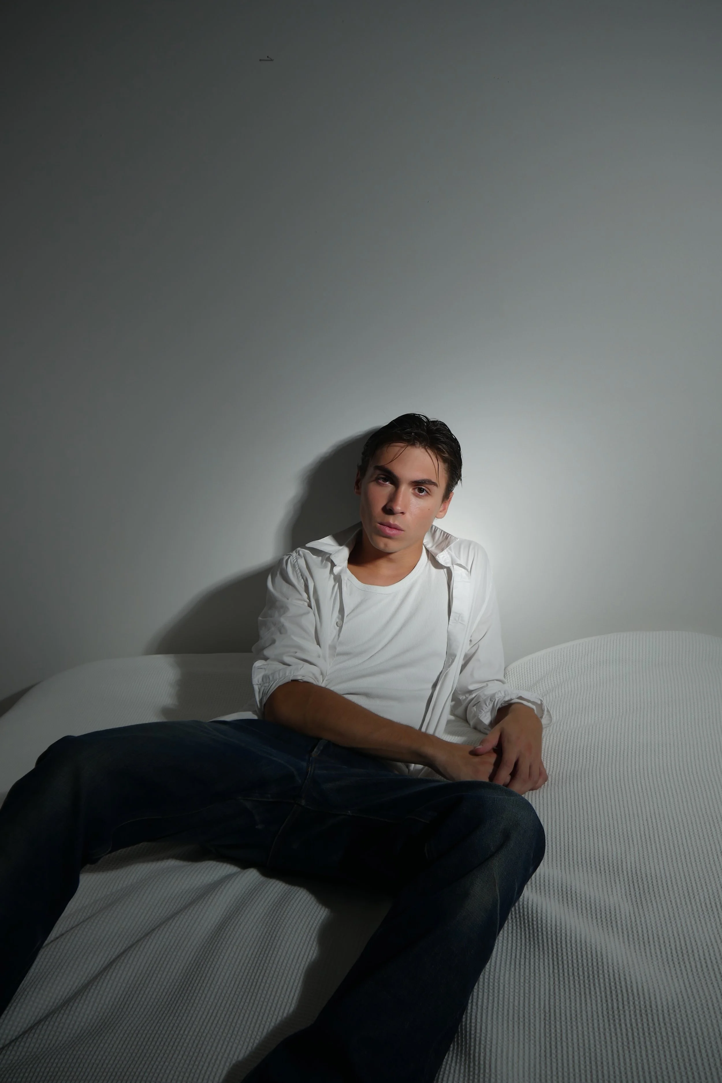 Un jeune homme assis sur un lit, portant une chemise blanche et un t-shirt, dans une pièce avec un mur gris clair.