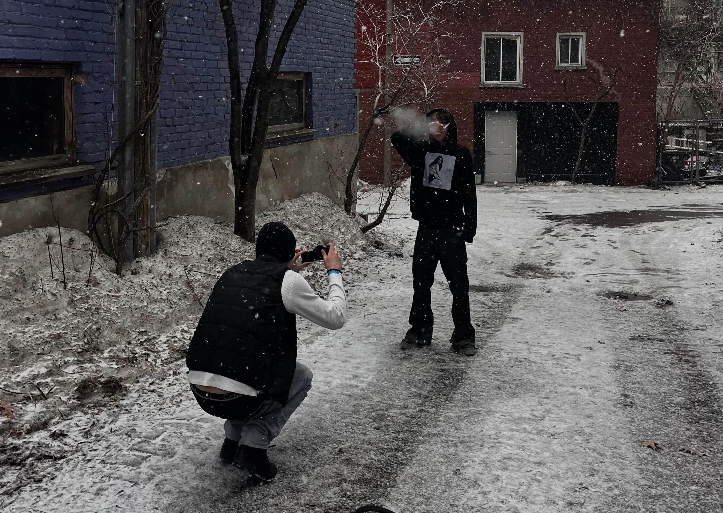 Deux jeunes hommes prennent une photo d'un autre homme qui fume une cigarette dehors sous la neige, devant des bâtiments colorés en bleu et rouge.