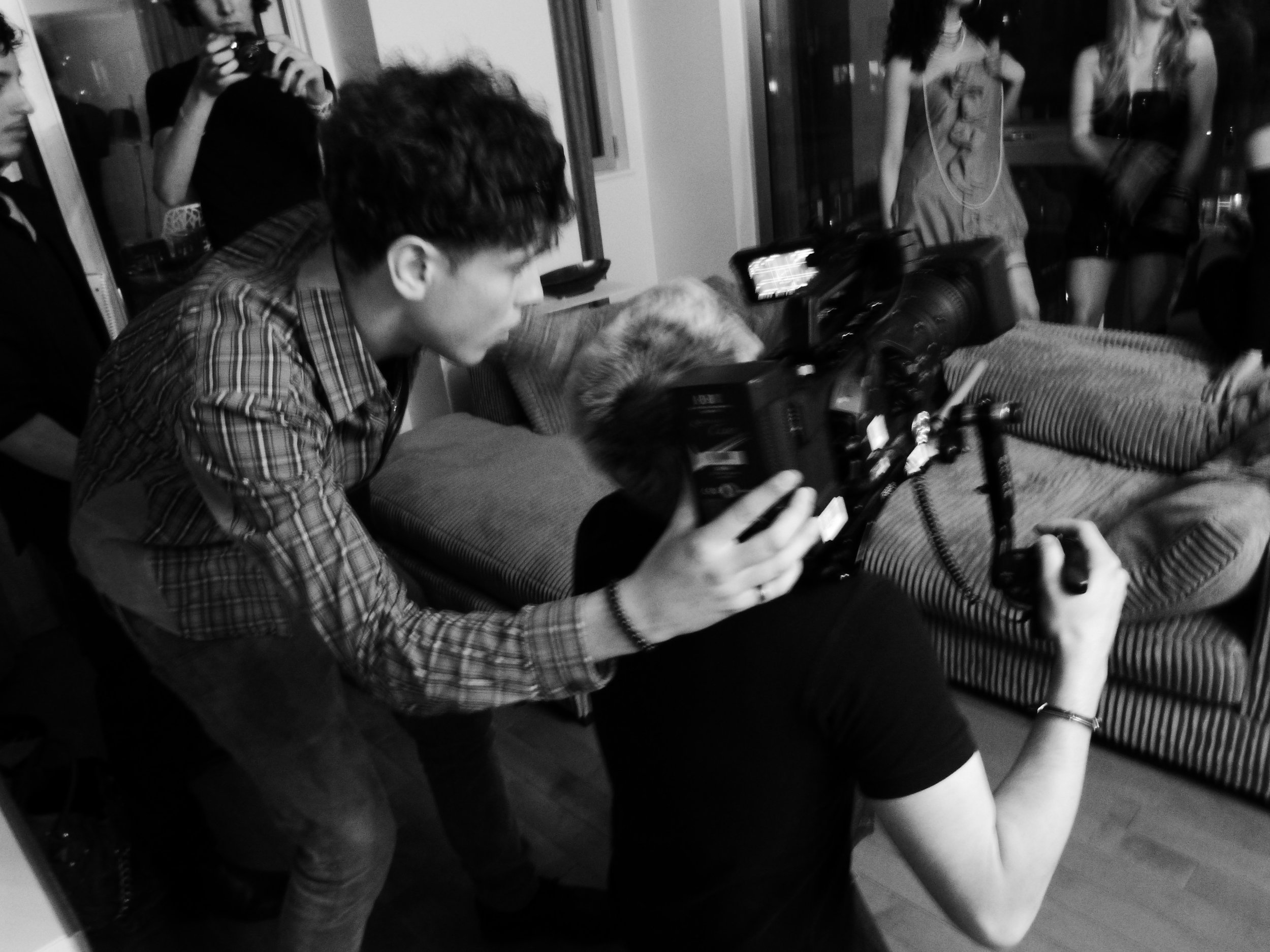Une scène en noir et blanc dans une pièce où plusieurs personnes, principalement des femmes, sont présentes. L'une d'entre elles est allongée sur un canapé ou un lit, tandis qu'une autre est en train de filmer ou de prendre une photo avec une caméra. D'autres personnes se tiennent autour, certaines avec des appareils électroniques ou des accessoires. L'ambiance semble détendue ou artistique.