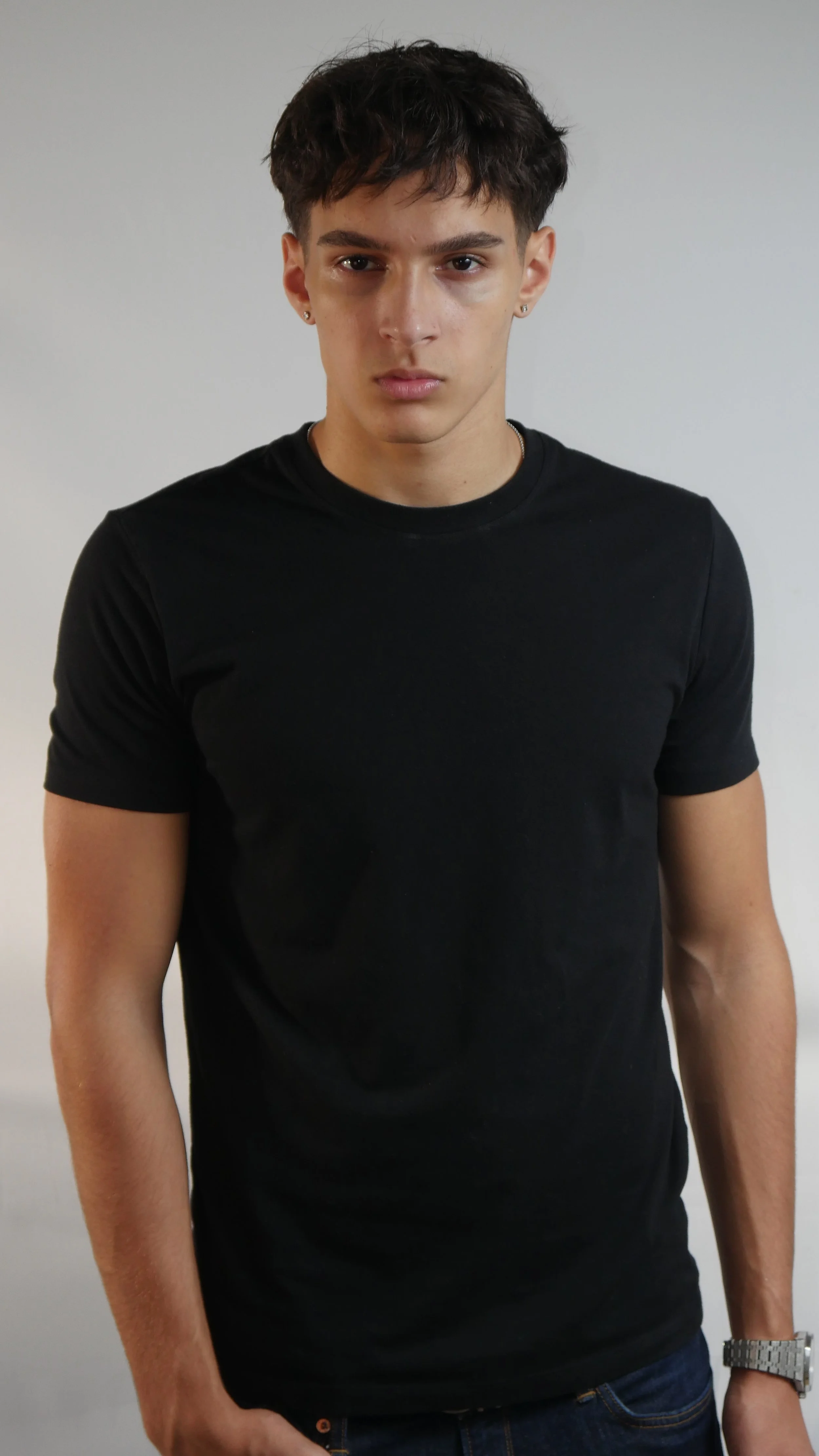 Jeune homme portant un t-shirt noir, avec le bras droit légèrement levé, regardant vers l'appareil photo, portant une montre et une boucle d'oreille.