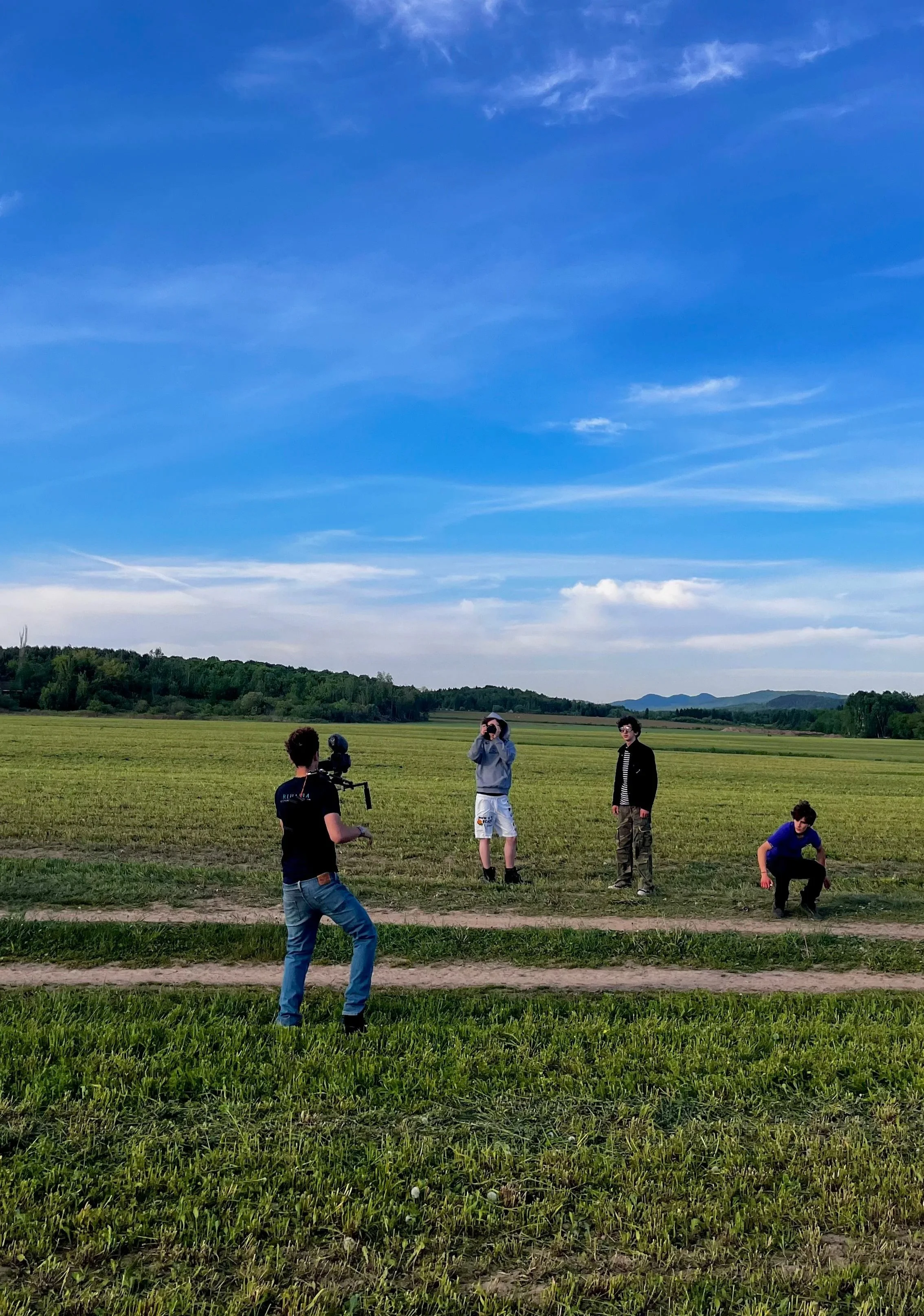 Un groupe de jeunes hommes dans un champ vert, certains avec du matériel photographique, sous un ciel bleu clair avec quelques nuages.