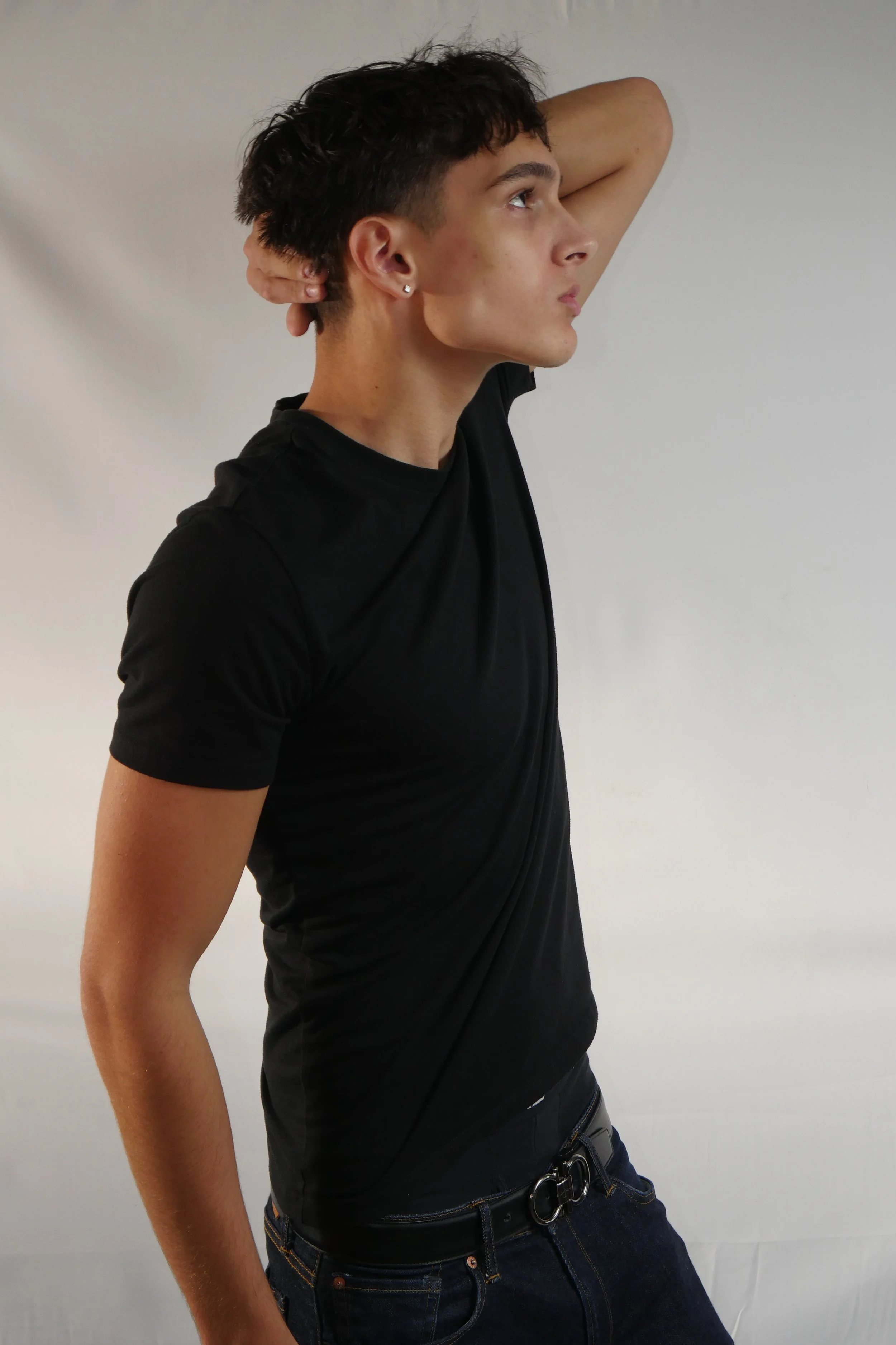 Jeune homme en t-shirt noir, posé de profil, se tenant la tête avec la main derrière le cou, fond neutre.