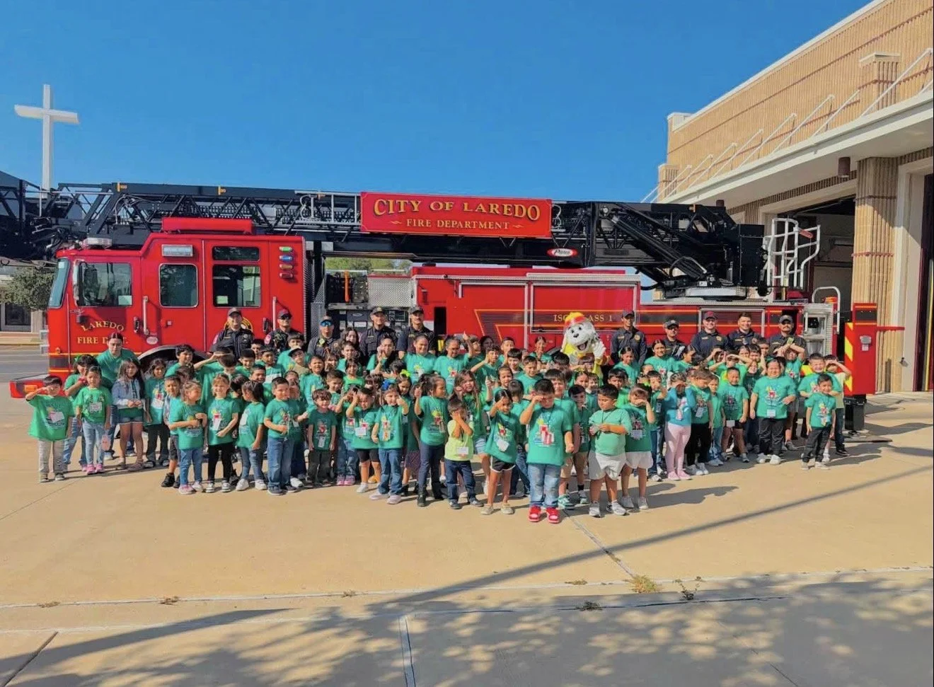 A group of children and adults gathered in front of a red fire truck with a ladder, on a sunny day outside a fire station. The children are wearing teal shirts, and some are making the peace sign or salutes, with a person in a firefighter costume and