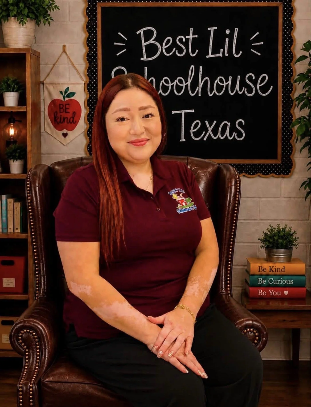 A woman with long red hair and light skin sitting in a leather armchair in a cozy room. She is smiling and wearing a maroon polo shirt with a colorful logo. Behind her, a chalkboard sign reads, 'Best Lil Schoolhouse Texas.' To her left, there is a bookshelf with potted plants and books, and a heart-shaped wall hanging that says, 'BE kind.' To her right, a small table has a potted plant and four colorful books with the titles, 'Be Kind,' 'Be Curious,' 'Be You,' and a fourth partially visible. The room has a warm, welcoming atmosphere.