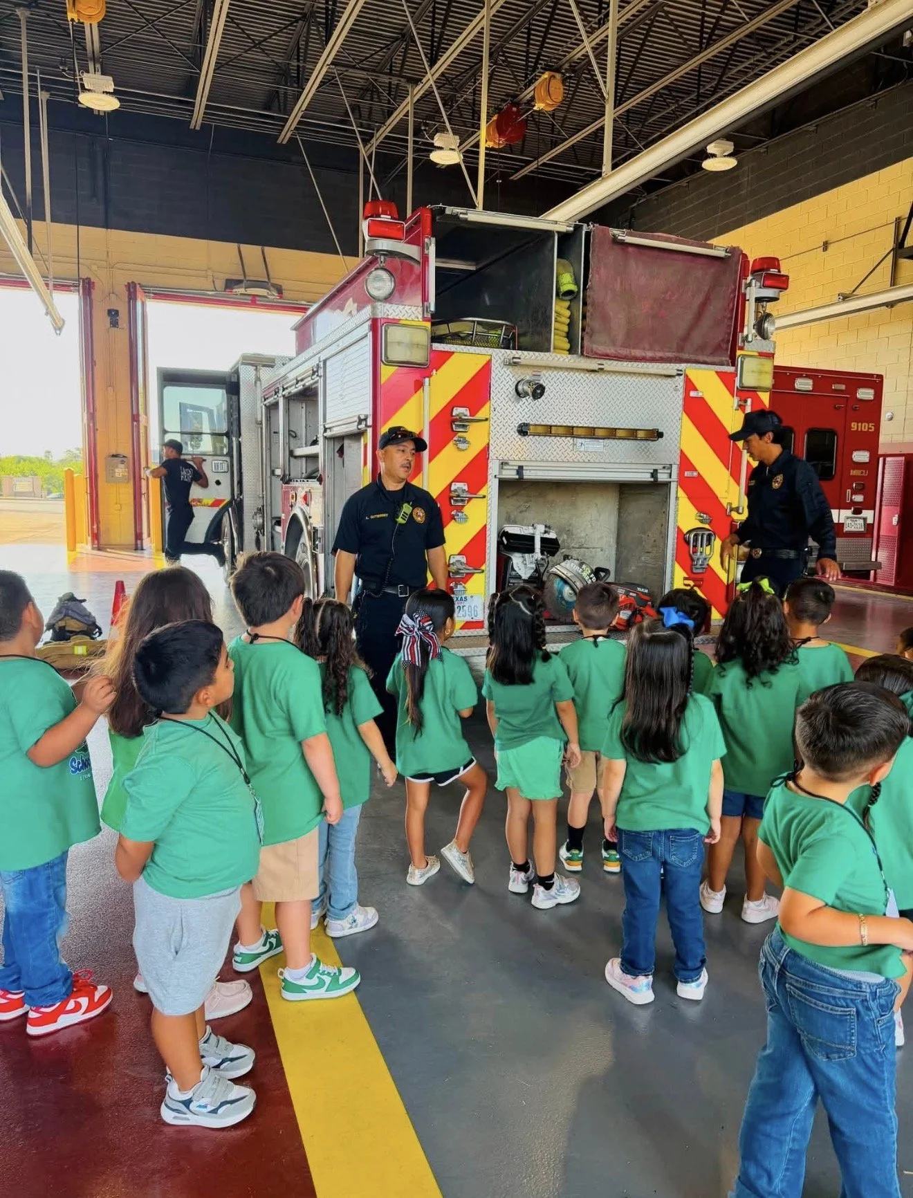 A group of young children in green shirts visiting a fire station. The children are listening to a firefighter in uniform who is explaining fire safety. A fire truck is parked behind them with open compartments showing equipment. The indoor area is w