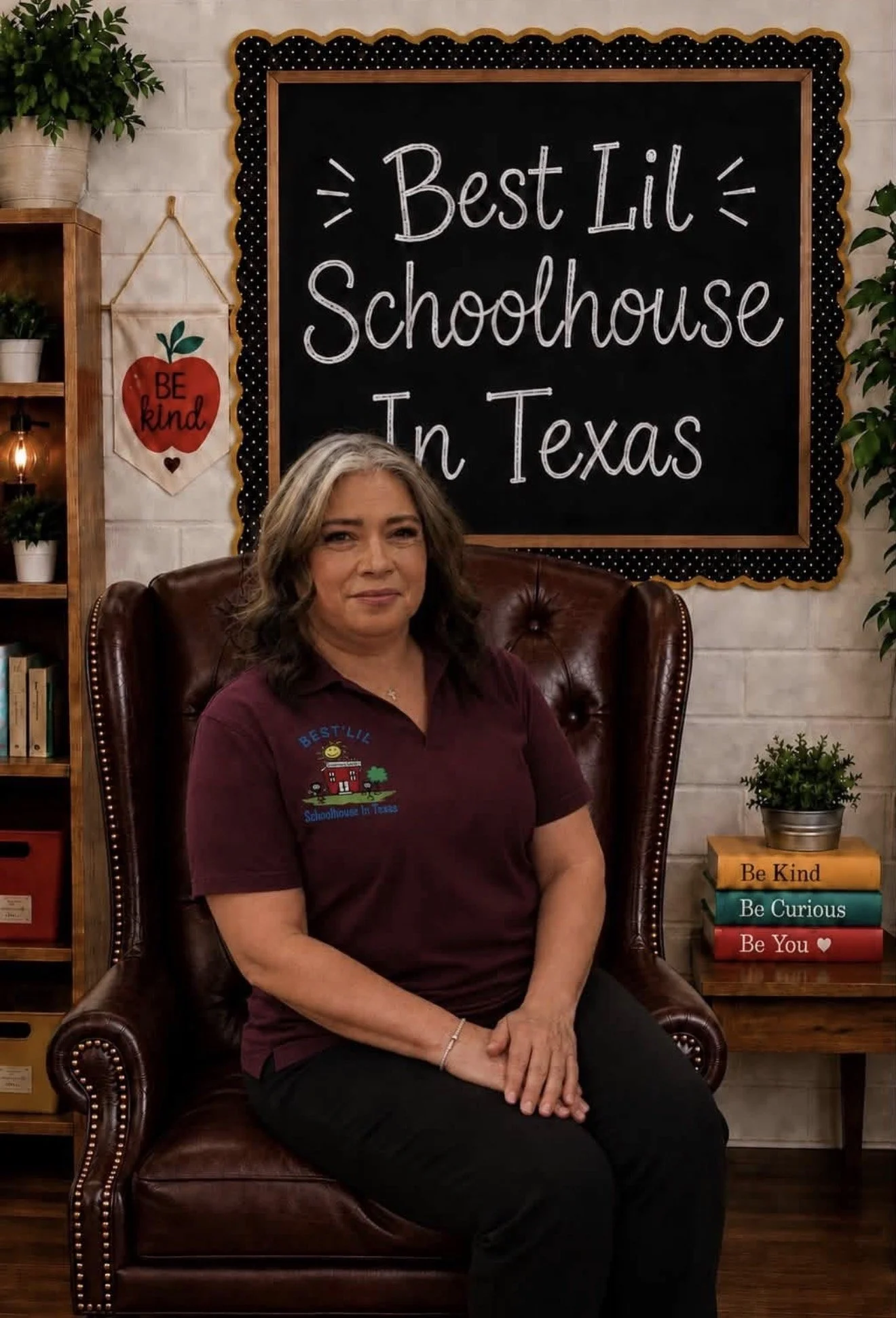 A woman with gray hair sitting on a brown leather armchair in front of a blackboard that reads 'Best Lil Schoolhouse In Texas.' She is wearing a maroon shirt with a schoolhouse logo and the words 'Best Lil Schoolhouse In Texas.' Behind her, to her right, are some stacked books and a potted plant. To her left, there is a wooden shelf with more plants and books.