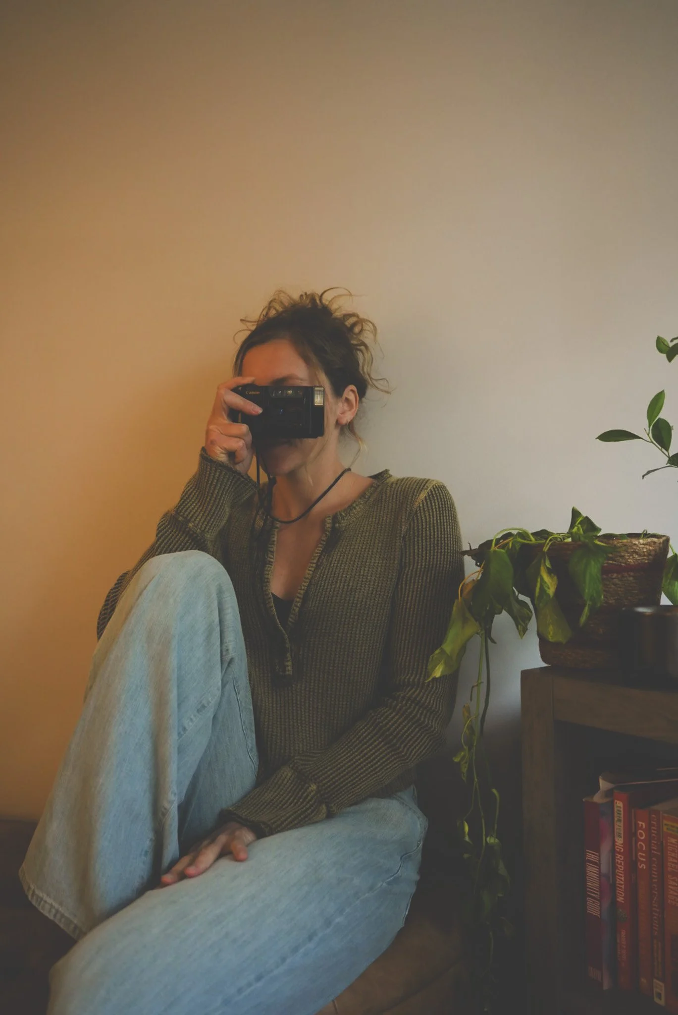 A person with curly hair taking a photo with a camera in a mirror, sitting with one leg crossed over the other, wearing a striped long-sleeve shirt and light-colored pants, with a potted plant and books on a shelf in the background.