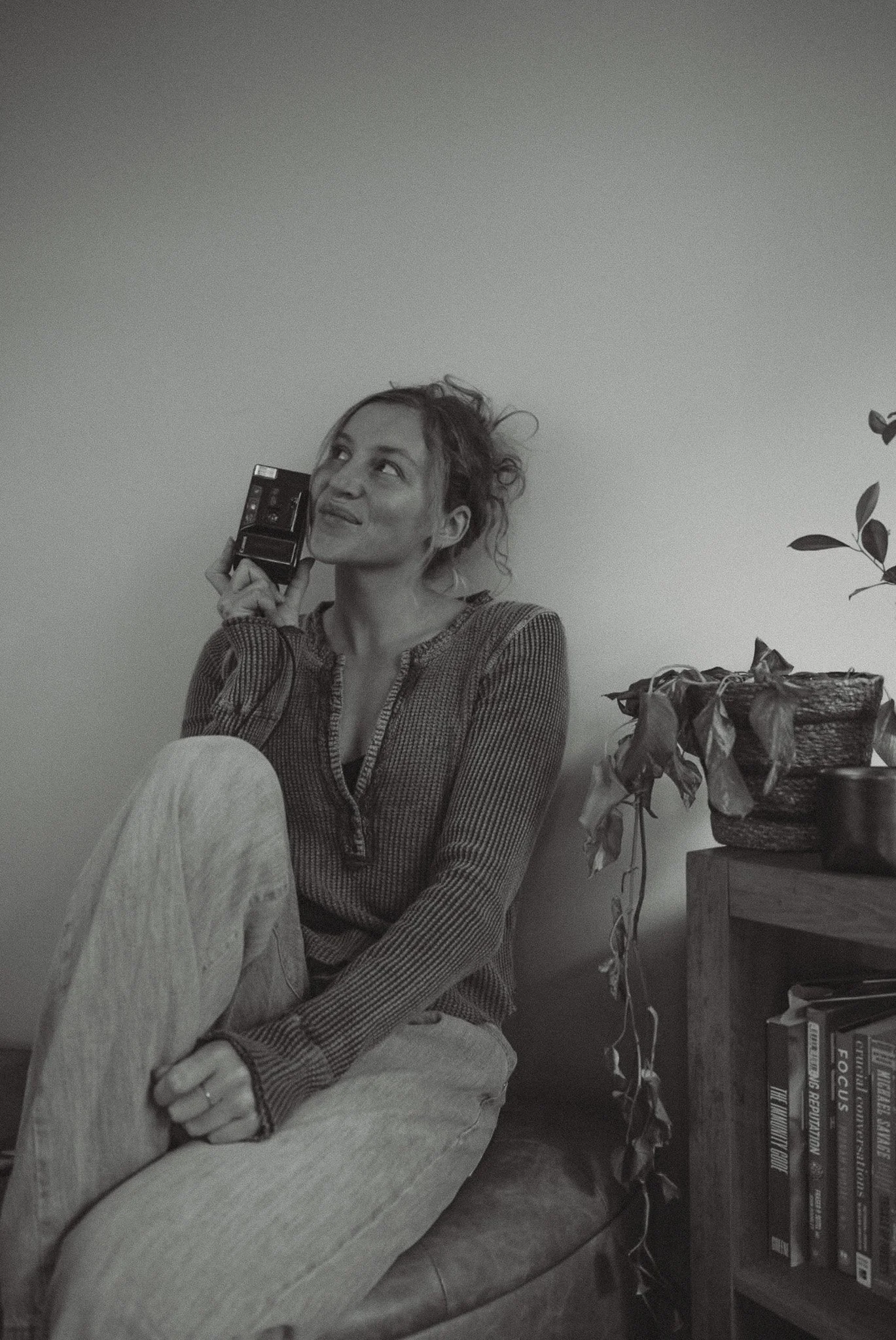 A woman with curly hair sitting on a chair, holding a phone to her ear, wearing a striped shirt and light-colored pants, with a potted plant and a bookshelf in the background.