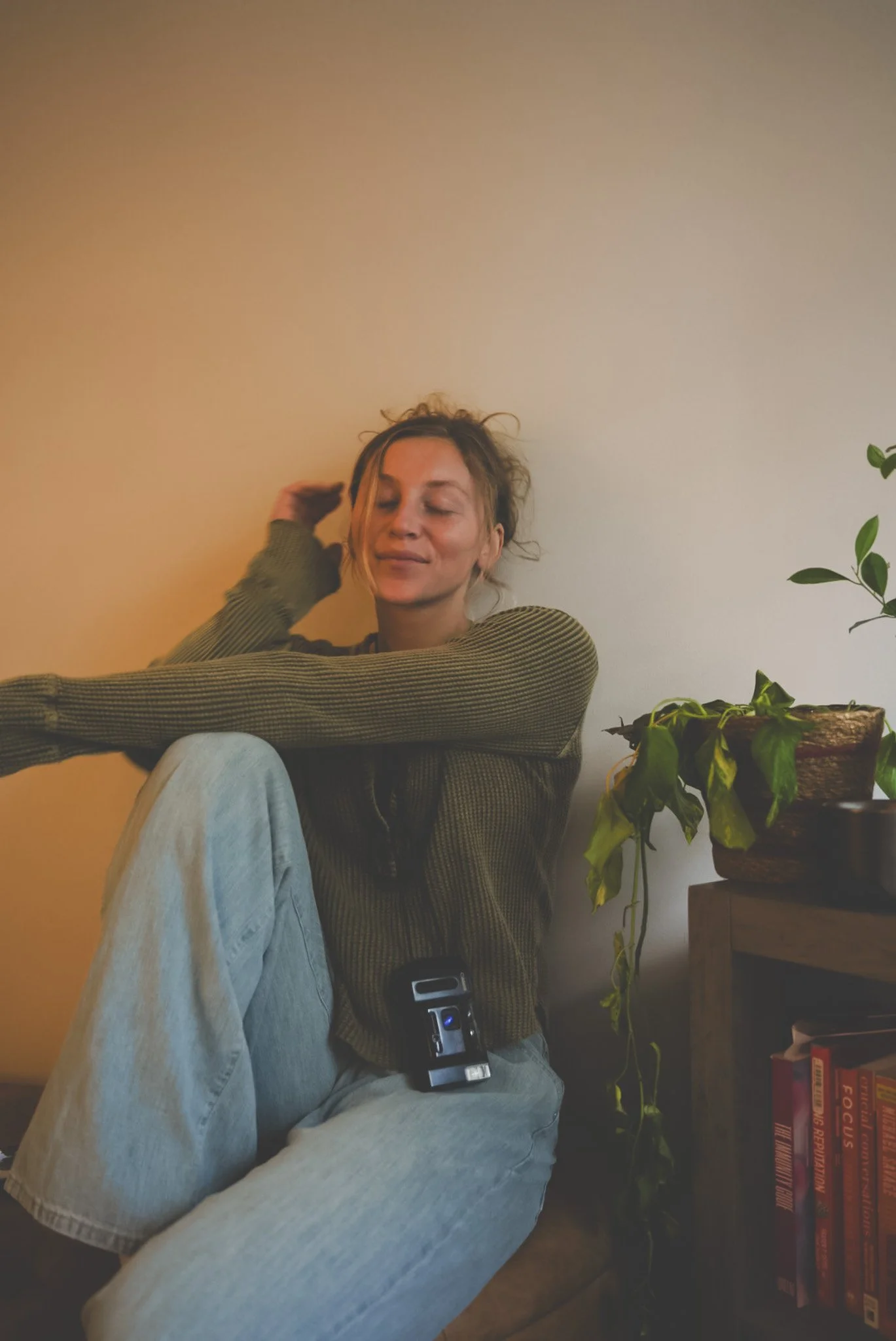 A woman with light brown hair and a gentle smile is sitting against a beige wall, wearing a green checkered shirt and light blue jeans. She has one hand resting on her head and appears relaxed.