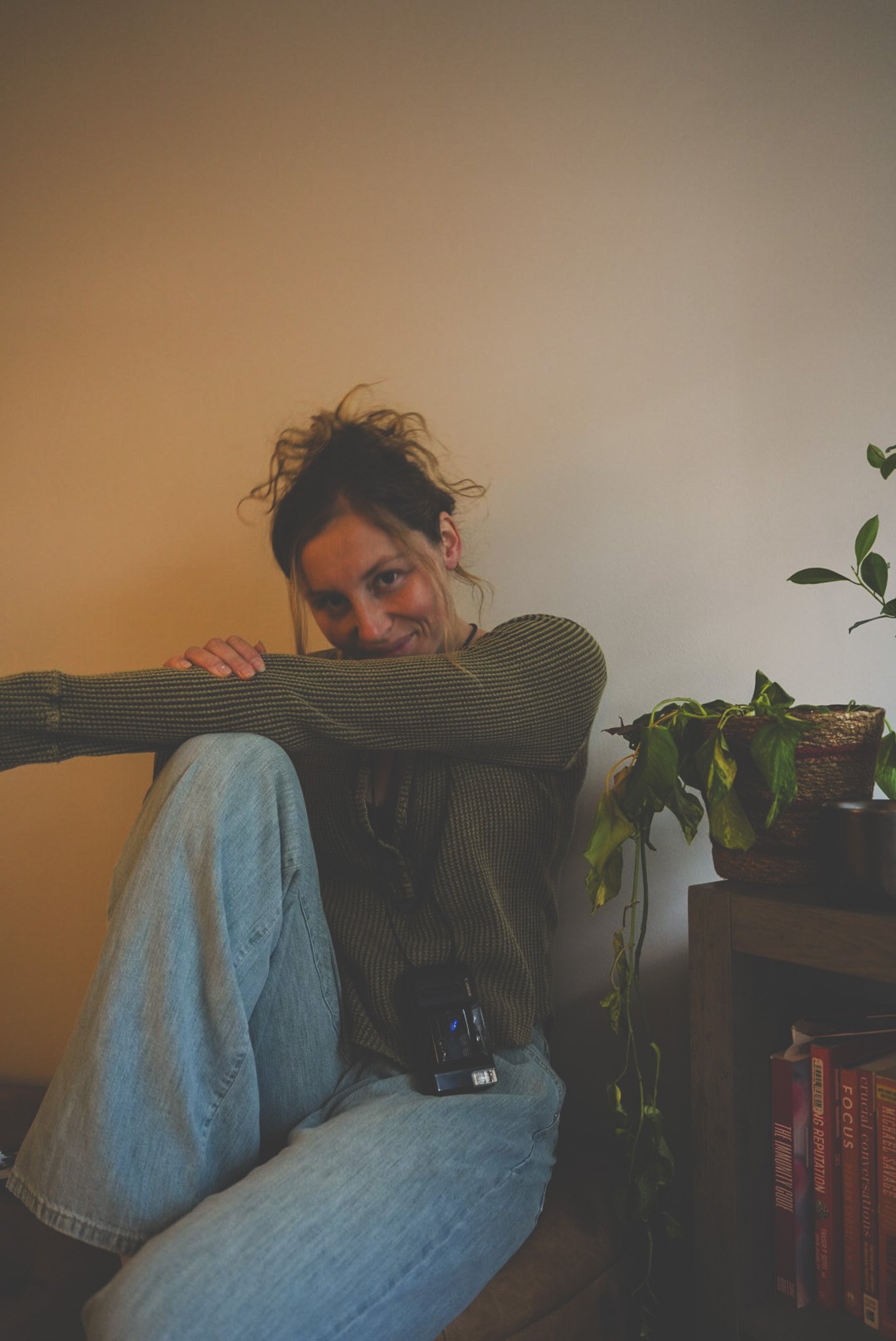 A woman with curly hair leaning on her knee and smiling at the camera, sitting in a room with a beige wall, a bookshelf with books, and a large potted plant.