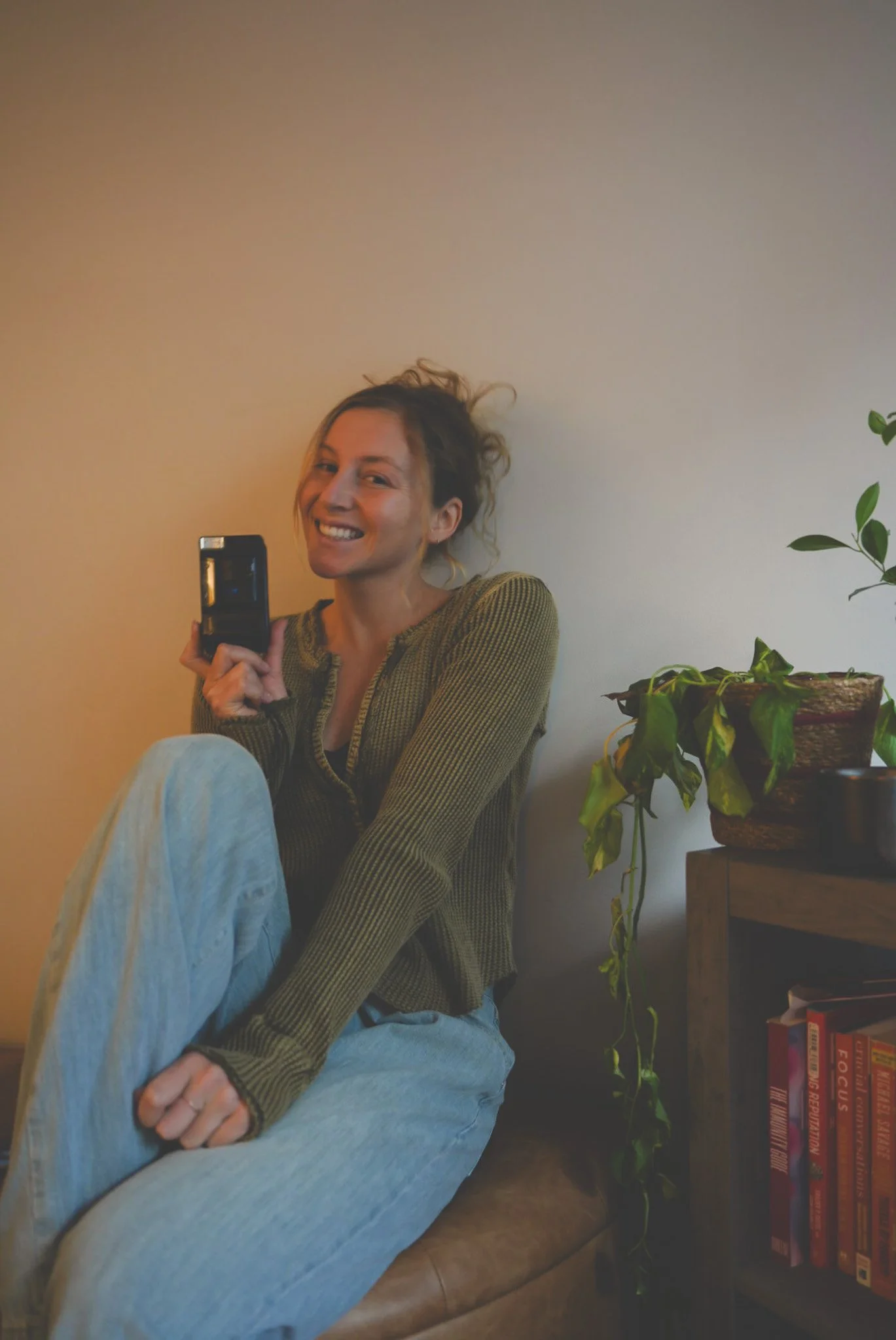 A woman with curly hair smiling, sitting on a couch, wearing a green sweater and light-colored pants, holding a phone, next to a potted plant and a bookshelf.