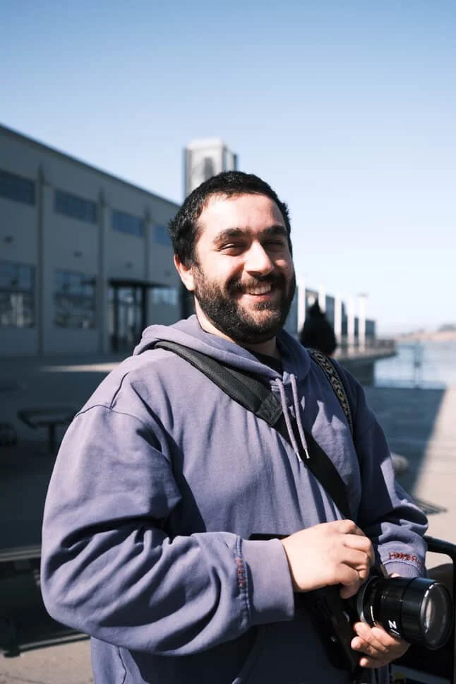 A smiling man with a beard in a purple hoodie holding a camera outdoors, with a building and water in the background on a sunny day.