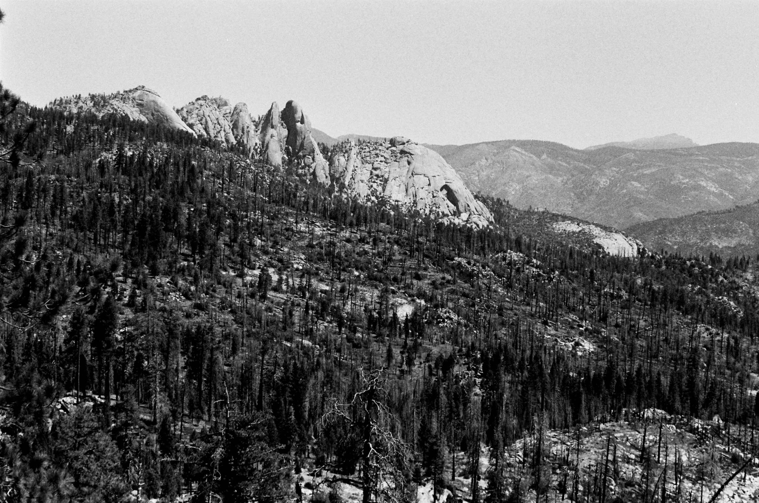 A black and white photograph of a mountainous landscape with rocky peaks, dense forested slopes, and distant mountains in the background.