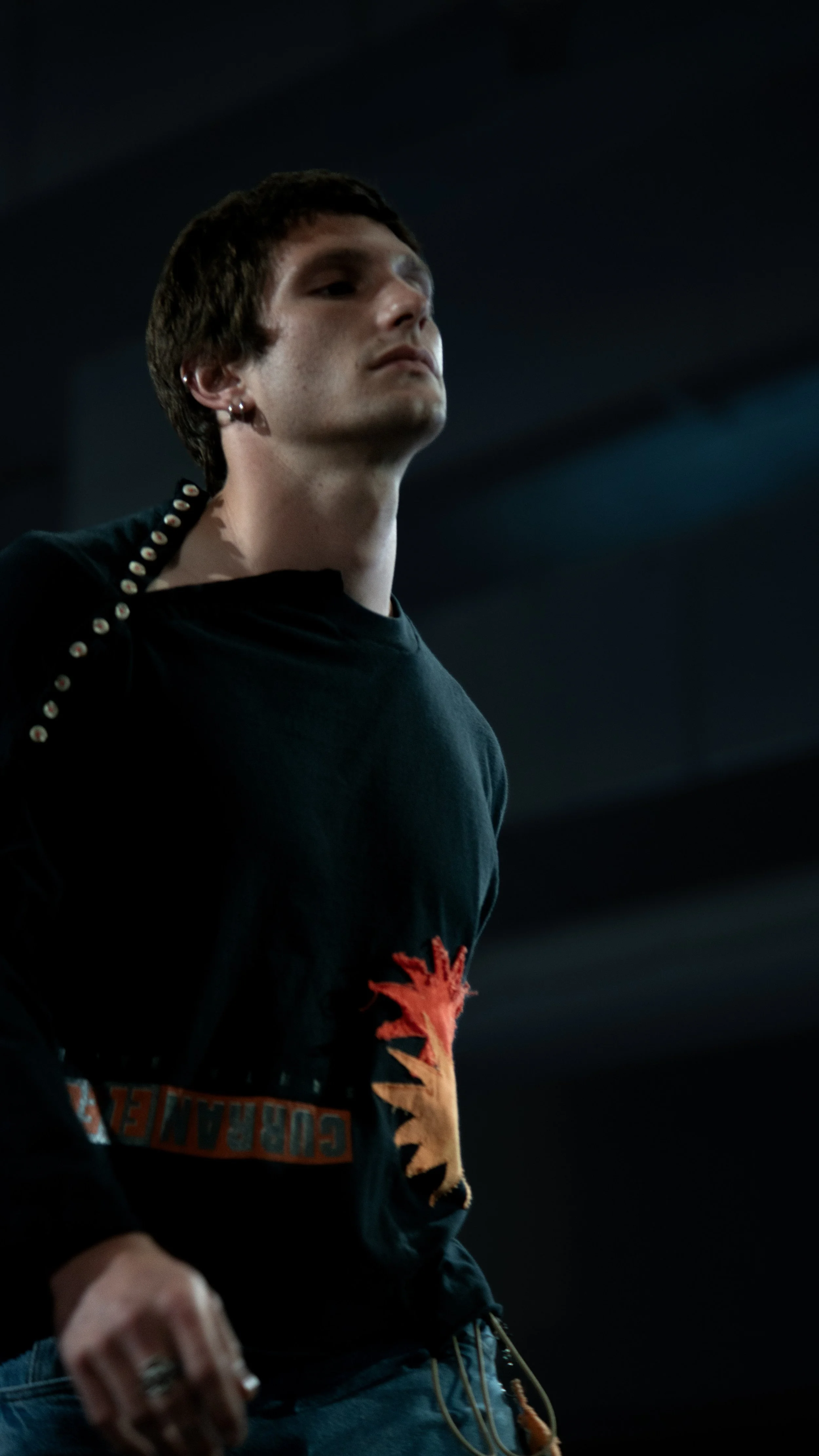 Close-up of a young person with short brown hair wearing a black hoodie with a red design and jeans, standing in a dark environment.