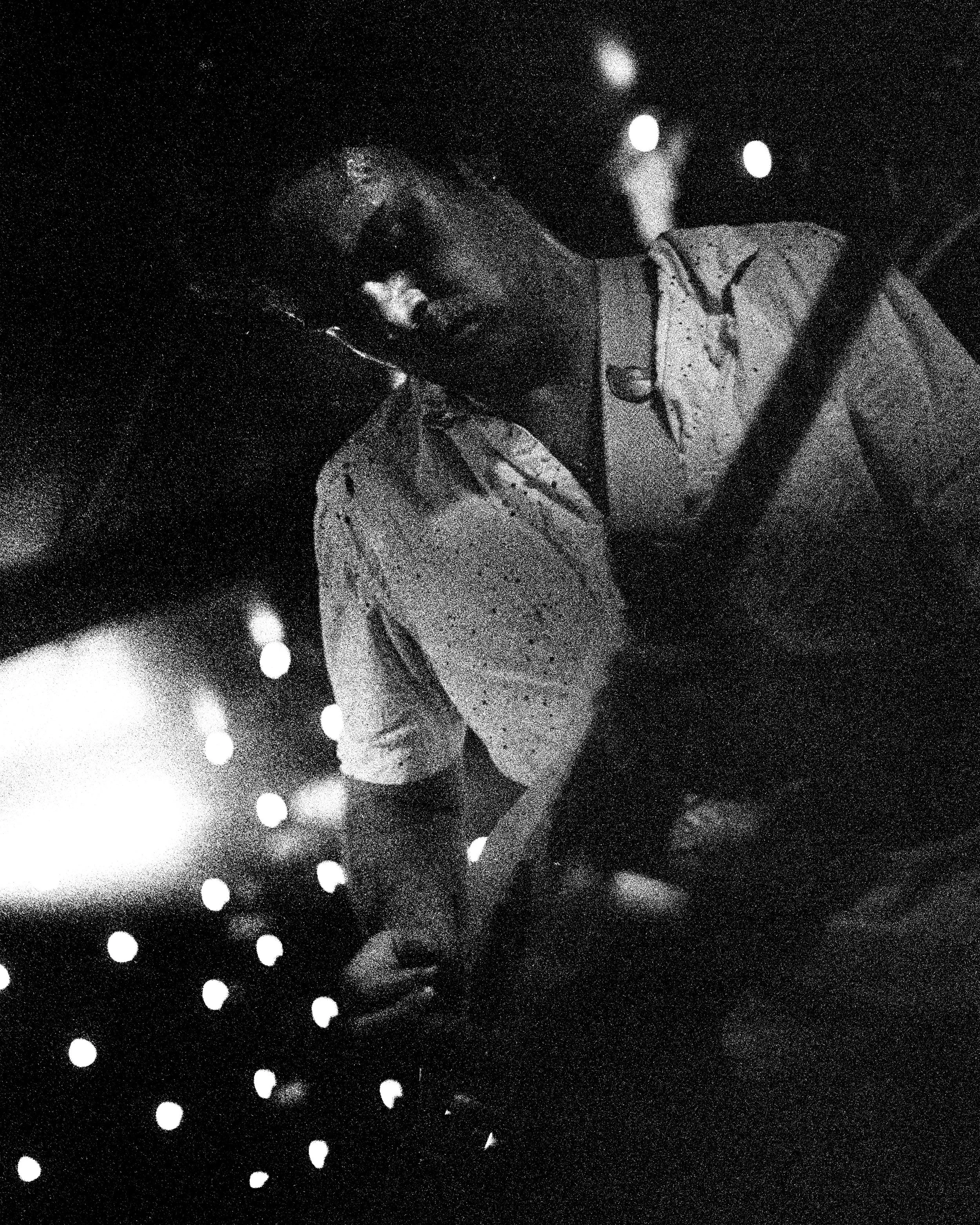 A person wearing glasses and a patterned shirt, with a backpack, appears to be in a dark, possibly indoor environment with bright lights in the background. The individual is looking downward and the photo has high contrast with visible noise.