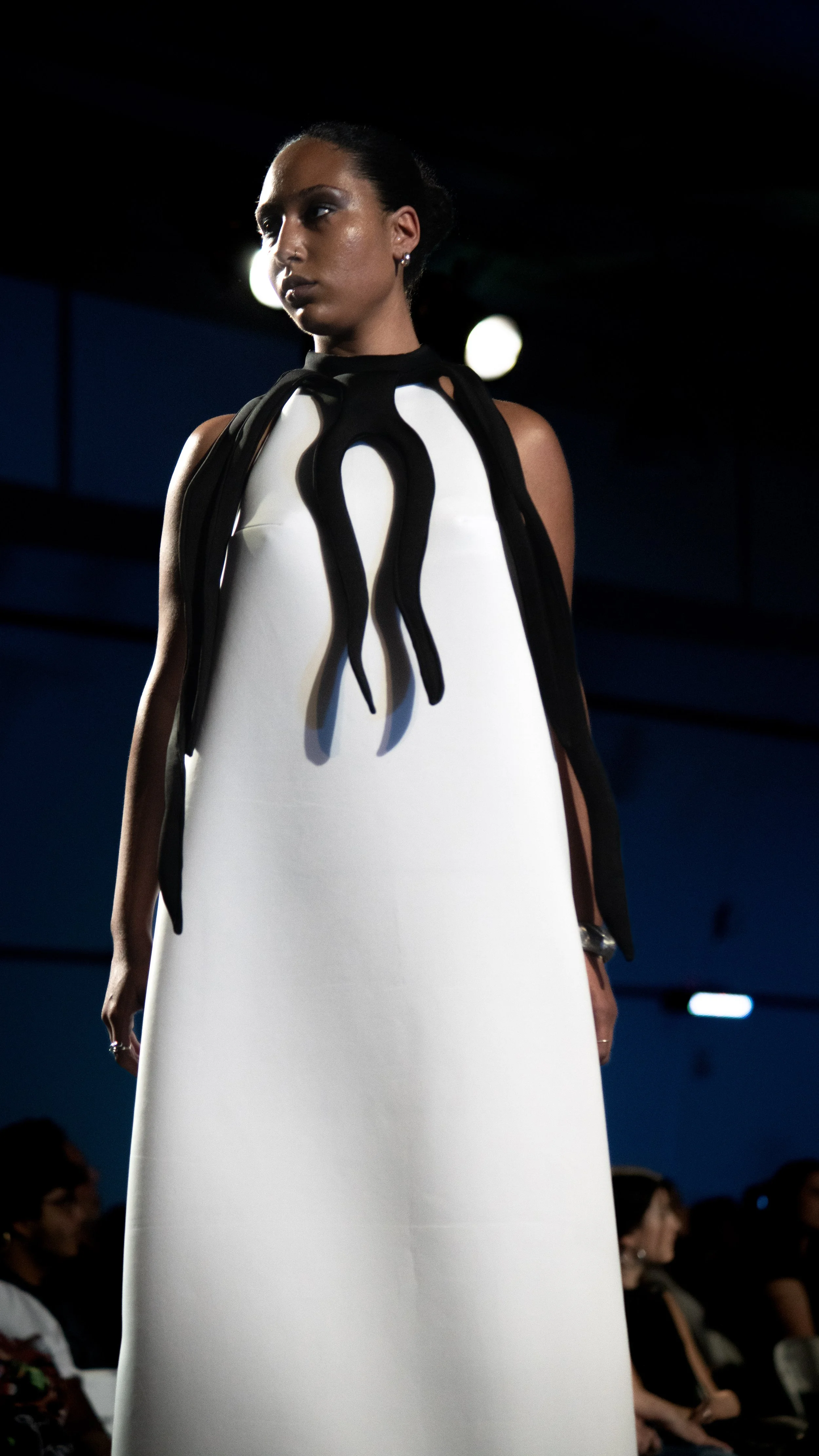 A woman with dark hair styled in a bun wearing a white dress with black design details, walking on a runway during a fashion show with an audience in the background.
