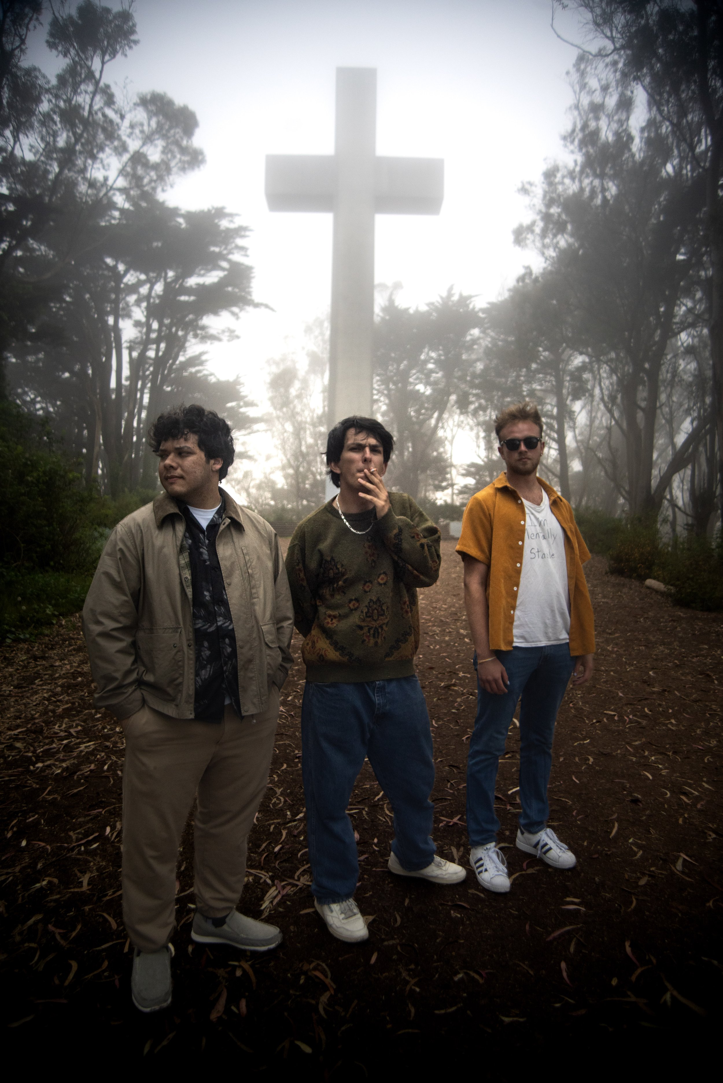 Three young men stand outdoors at sunset with a large cross and trees in the background. They are dressed in casual clothing and appear relaxed.
