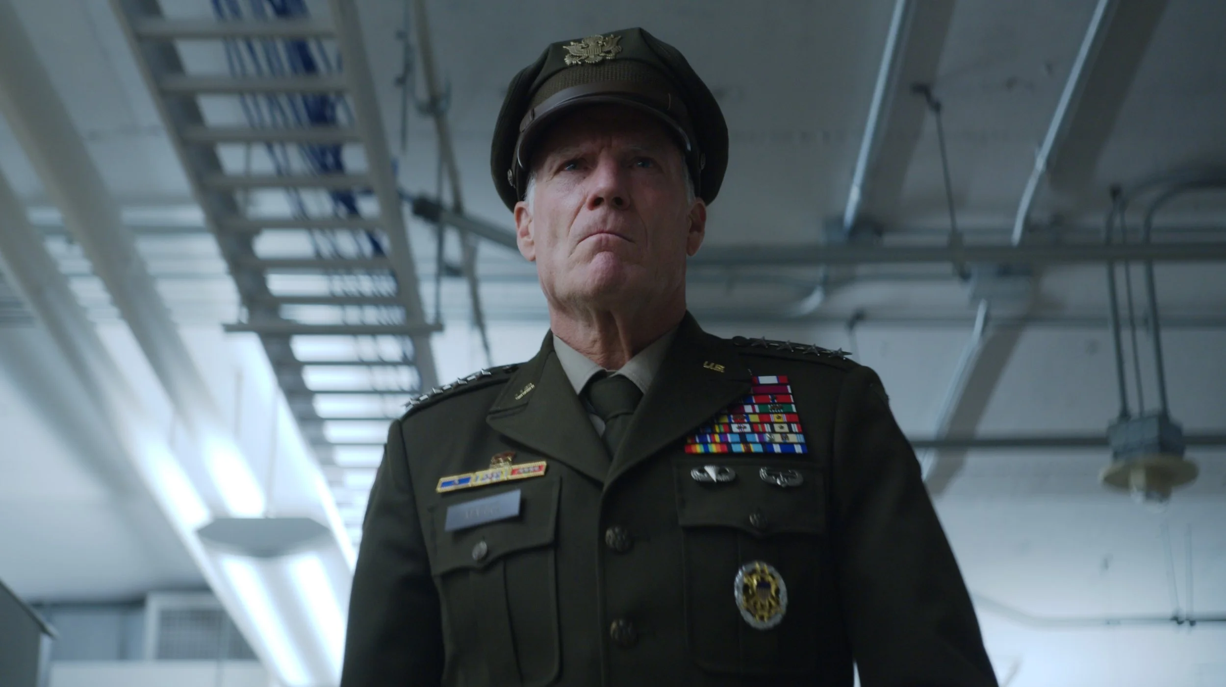 A senior military officer wearing uniform with medals and badges, and a peaked cap, standing in an industrial or military setting with metal staircases in the background.