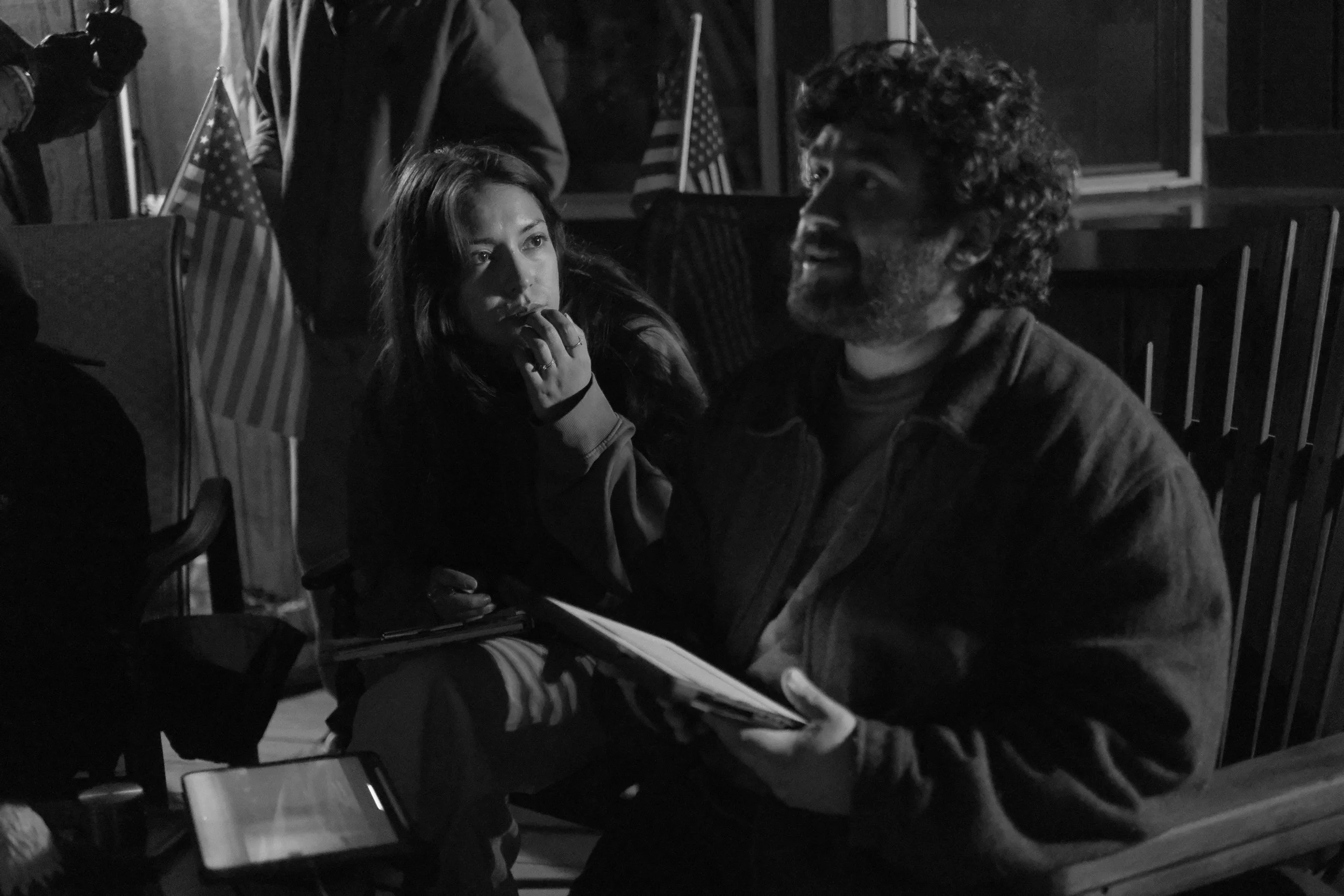 A black-and-white photo of two people sitting at a table, engaged in conversation. The woman on the left has long hair, is resting her chin on her hand, and appears attentive. The man on the right has curly hair and a beard, is smiling, and is holding a notebook. In the background, there are American flags and other individuals.