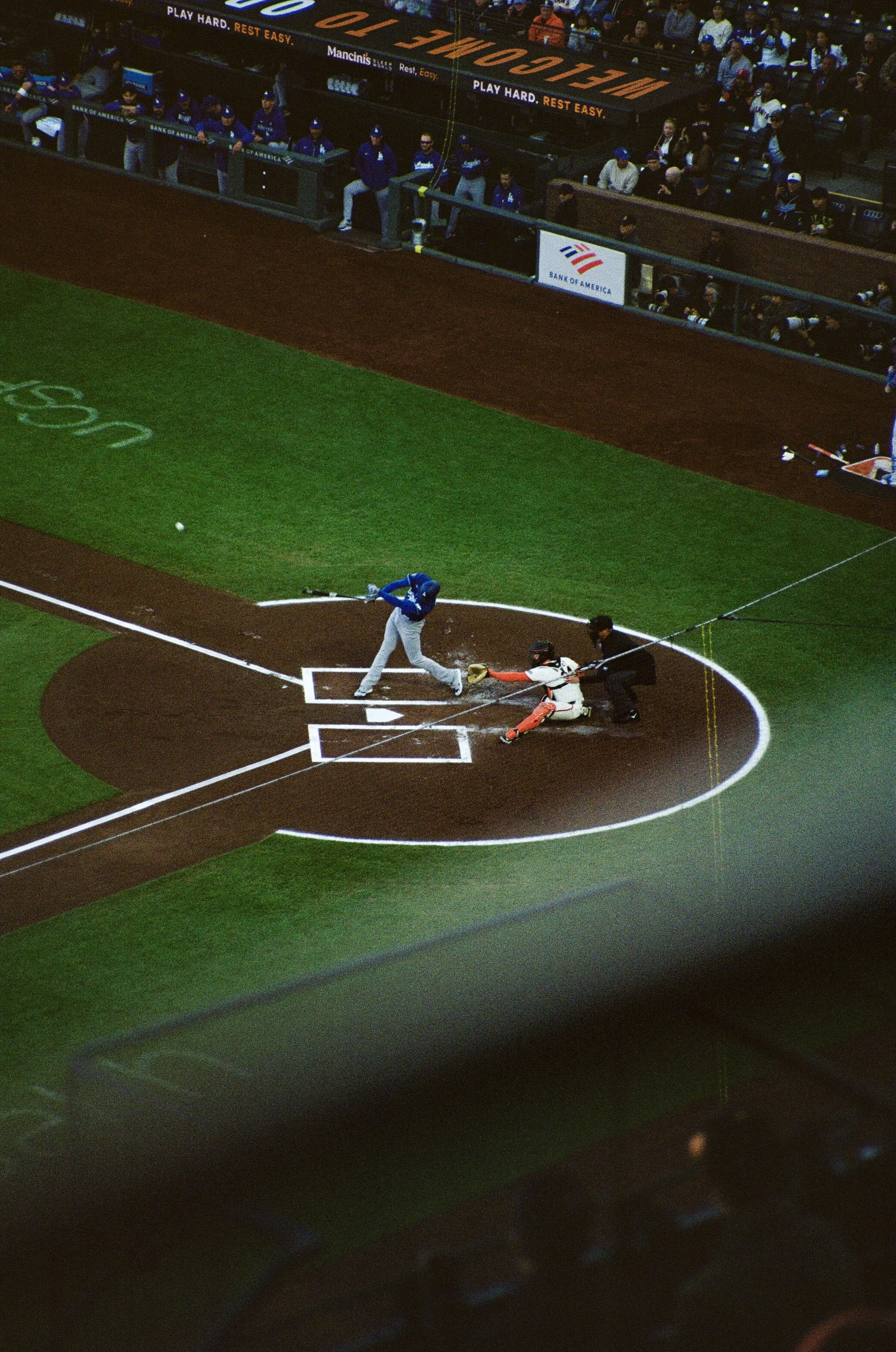 Baseball game with batter swinging at a pitch, catcher and umpire in position, and spectators in the stands.
