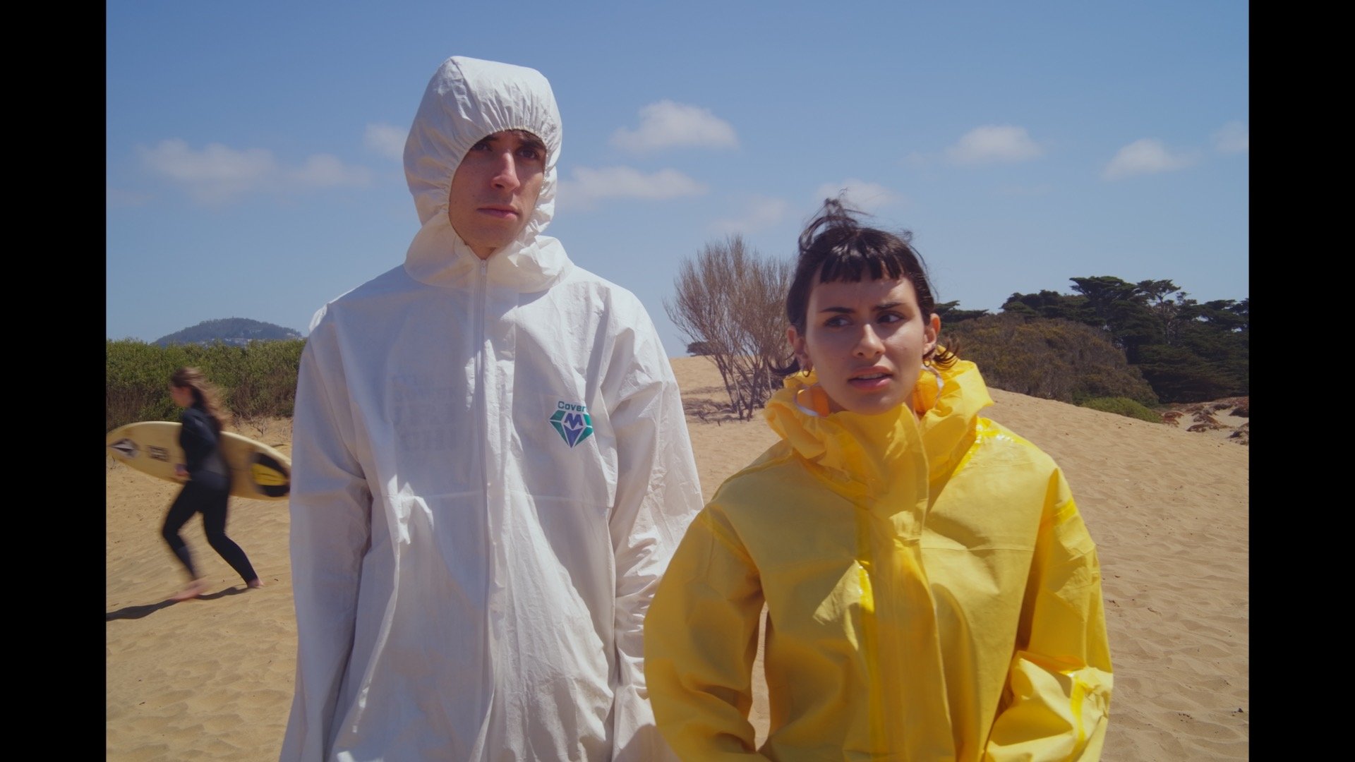 Two people on a beach, wearing protective rain gear, with a woman carrying a surfboard in the background.
