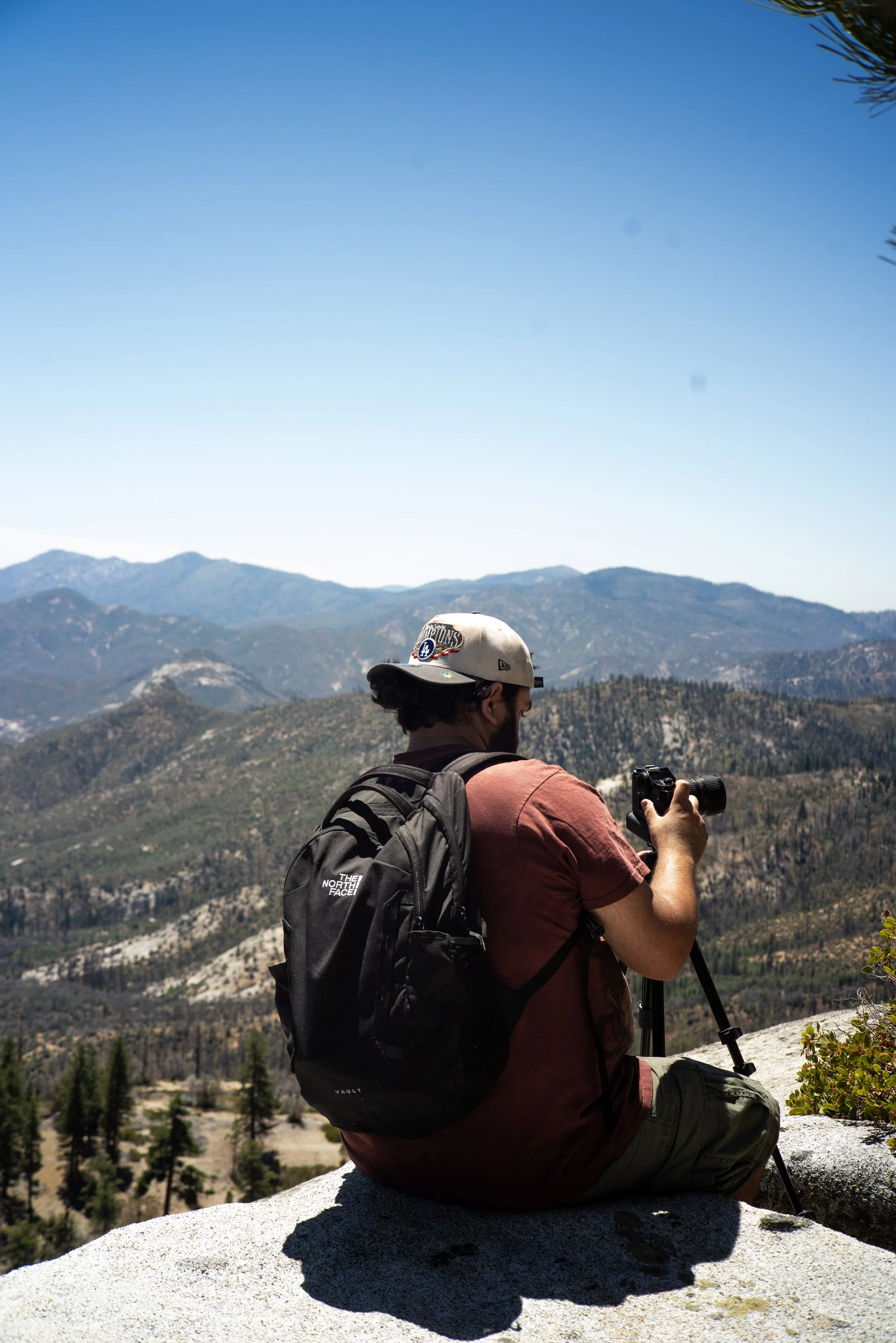 A man with a backpack sitting on a rock, holding a camera, overlooking a mountainous landscape with trees and rolling hills under a clear blue sky.