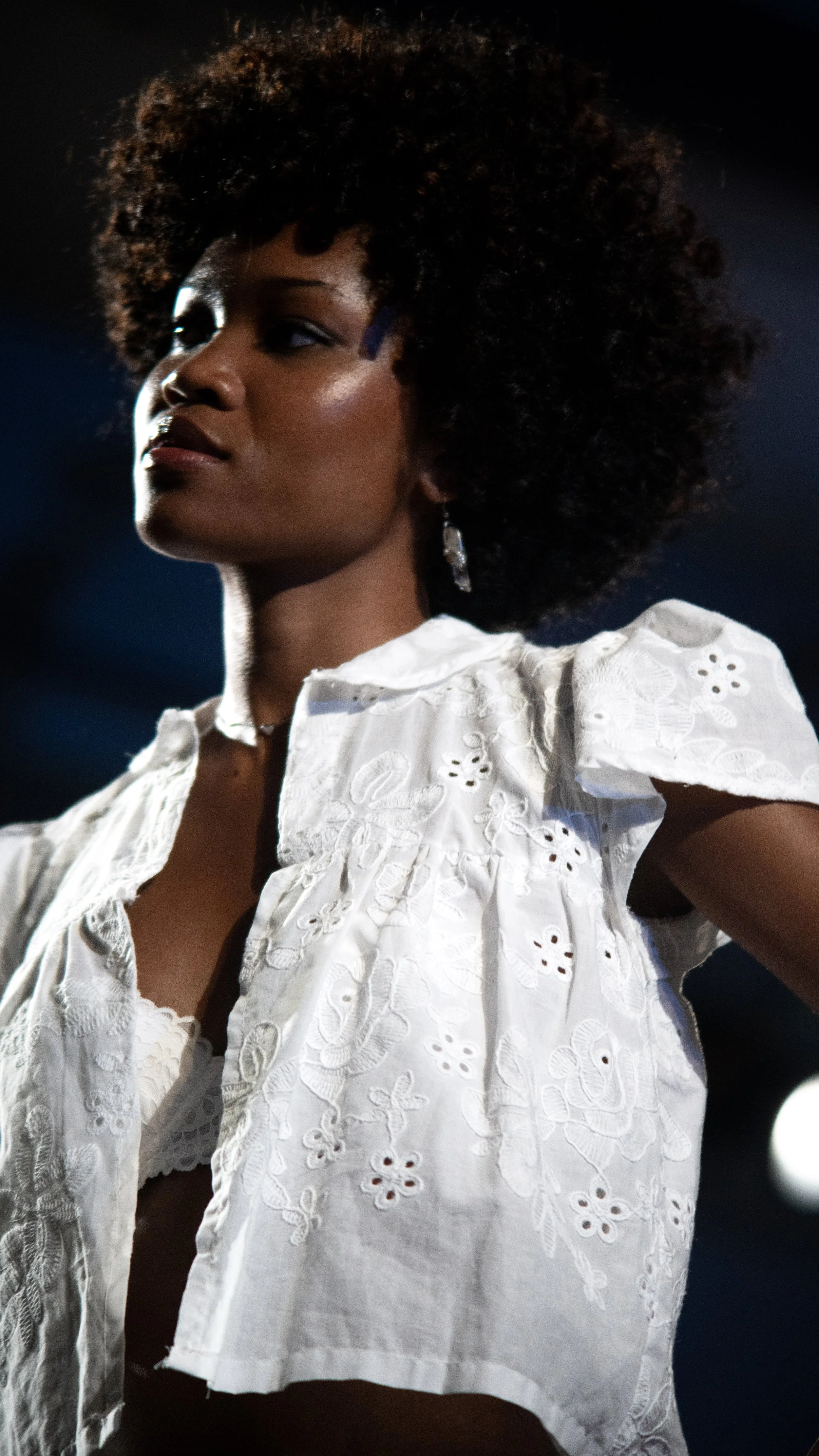Close-up of a woman with dark skin and curly hair wearing a white embroidered blouse and silver earrings, with stage lighting in the background.