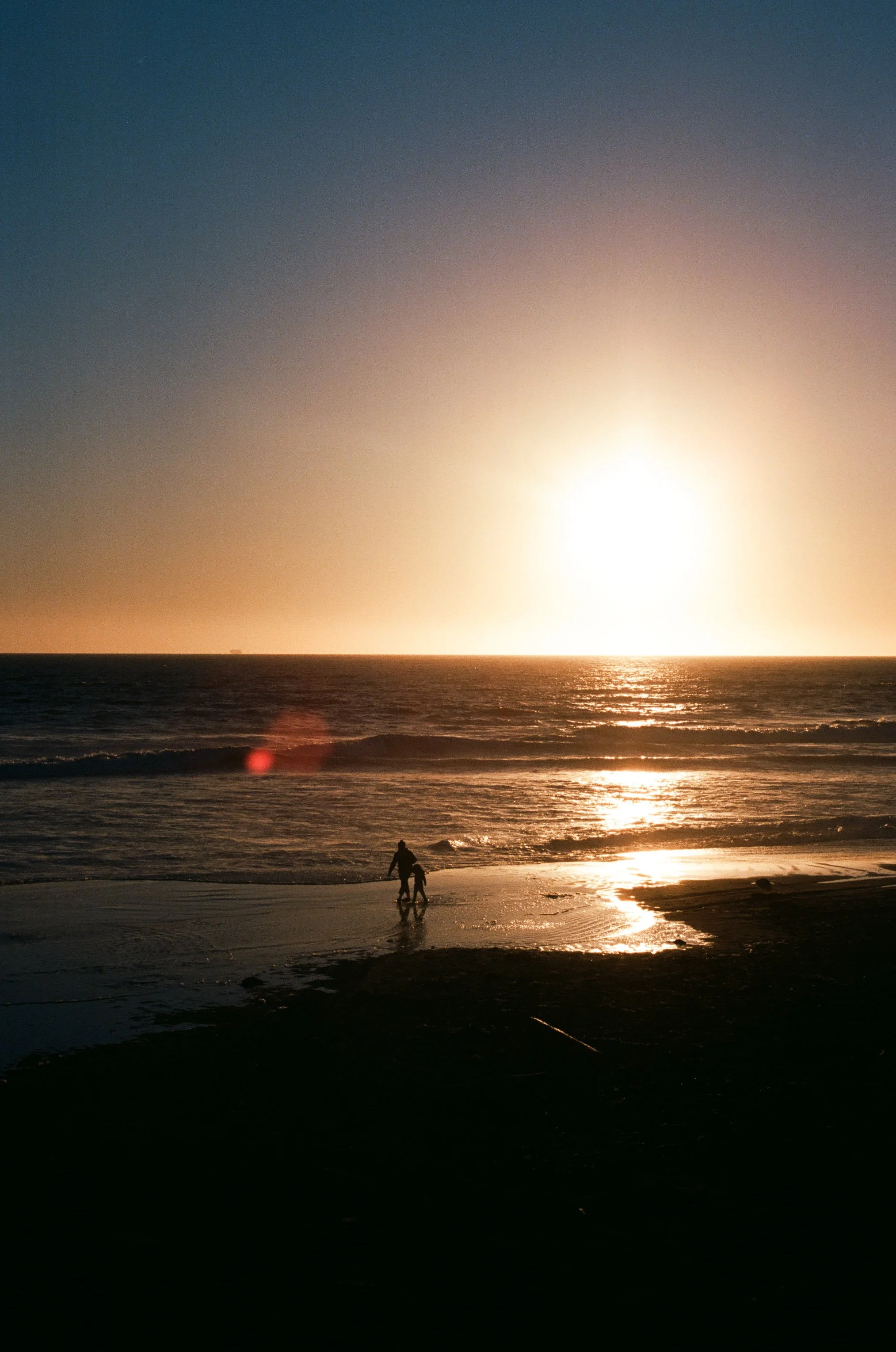 A sunset over the ocean with a person and a child walking along the beach.