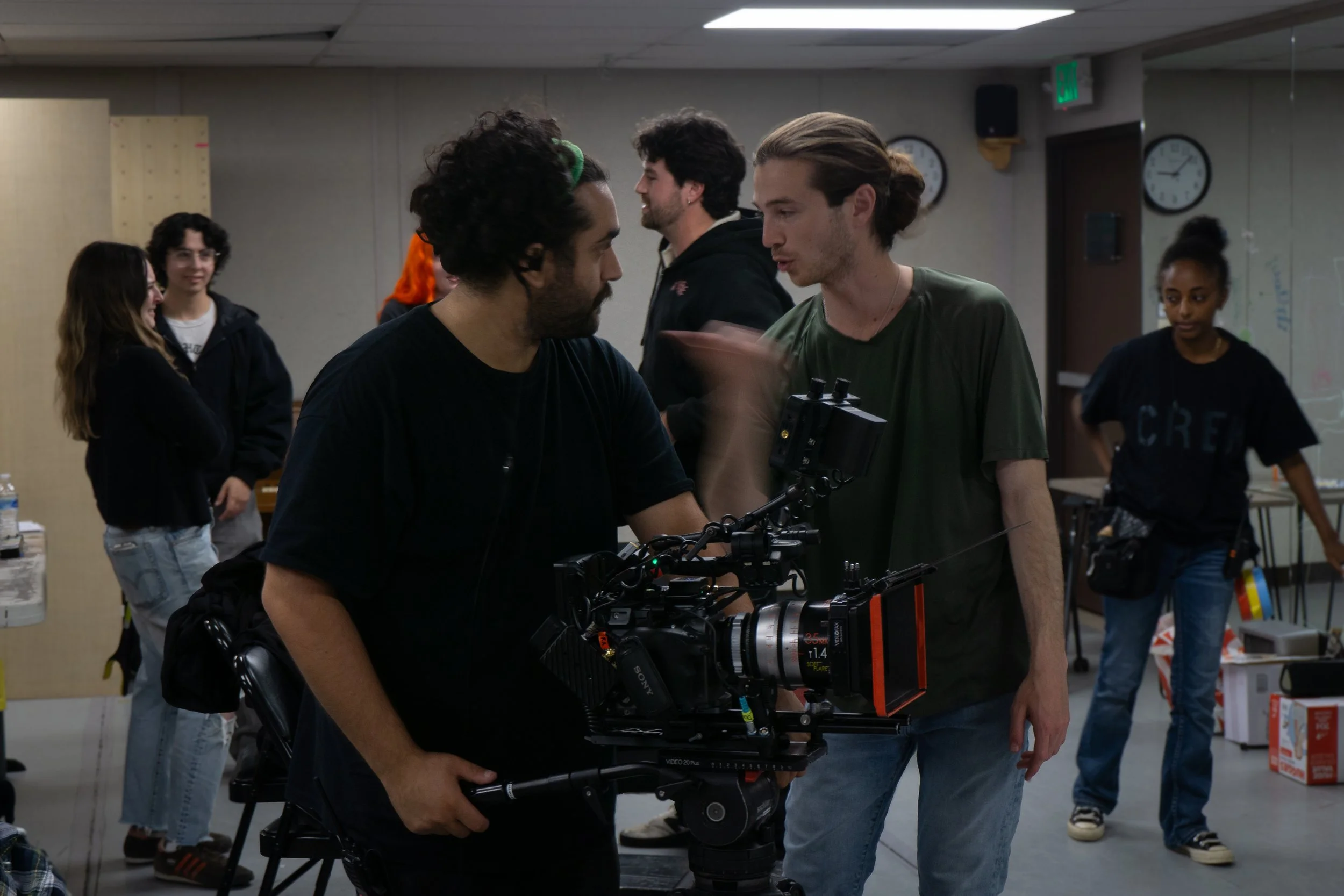 Filmmaker or camera operator adjusting camera equipment on a film set or production, with crew members in the background.