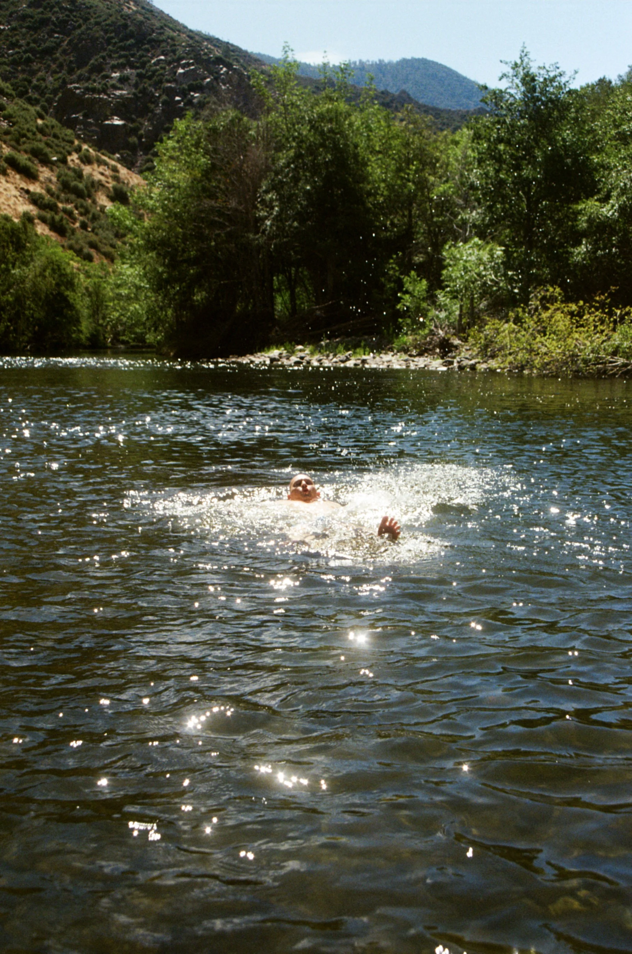 A person swimming in a river with forested hills and mountains in the background.