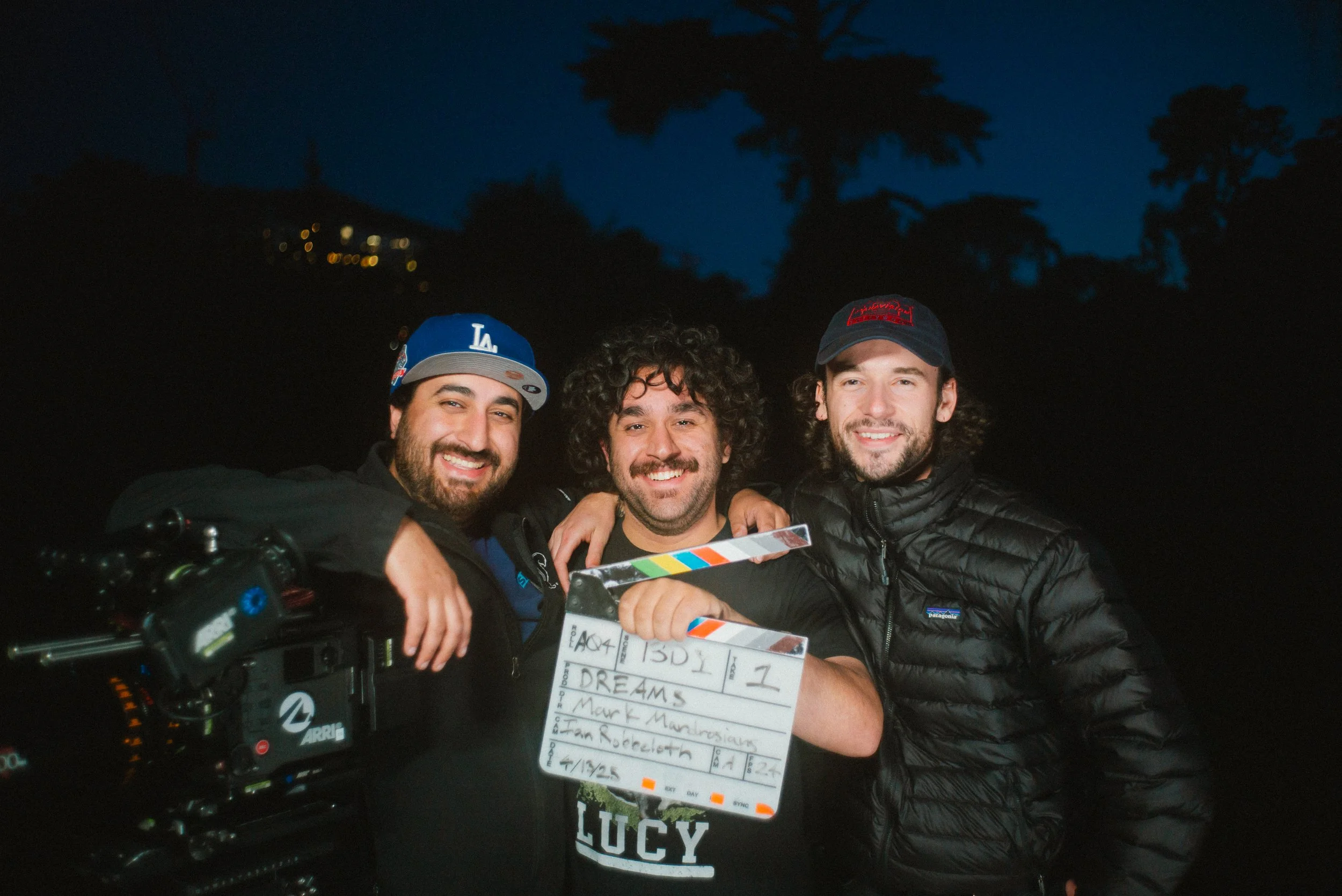 Three smiling men holding a clapperboard in front of a dark outdoor setting with trees and a few distant lights in the background.