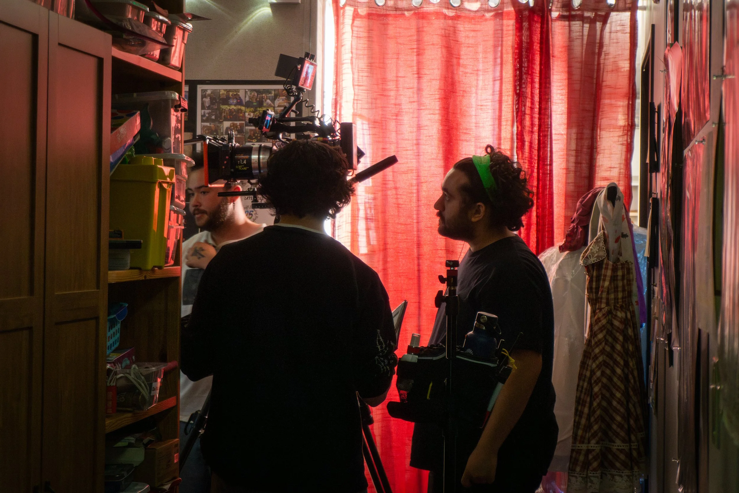 Three people in a room with red curtains, surrounded by various equipment, including cameras and a computer setup, appear to be engaged in filming or photography.