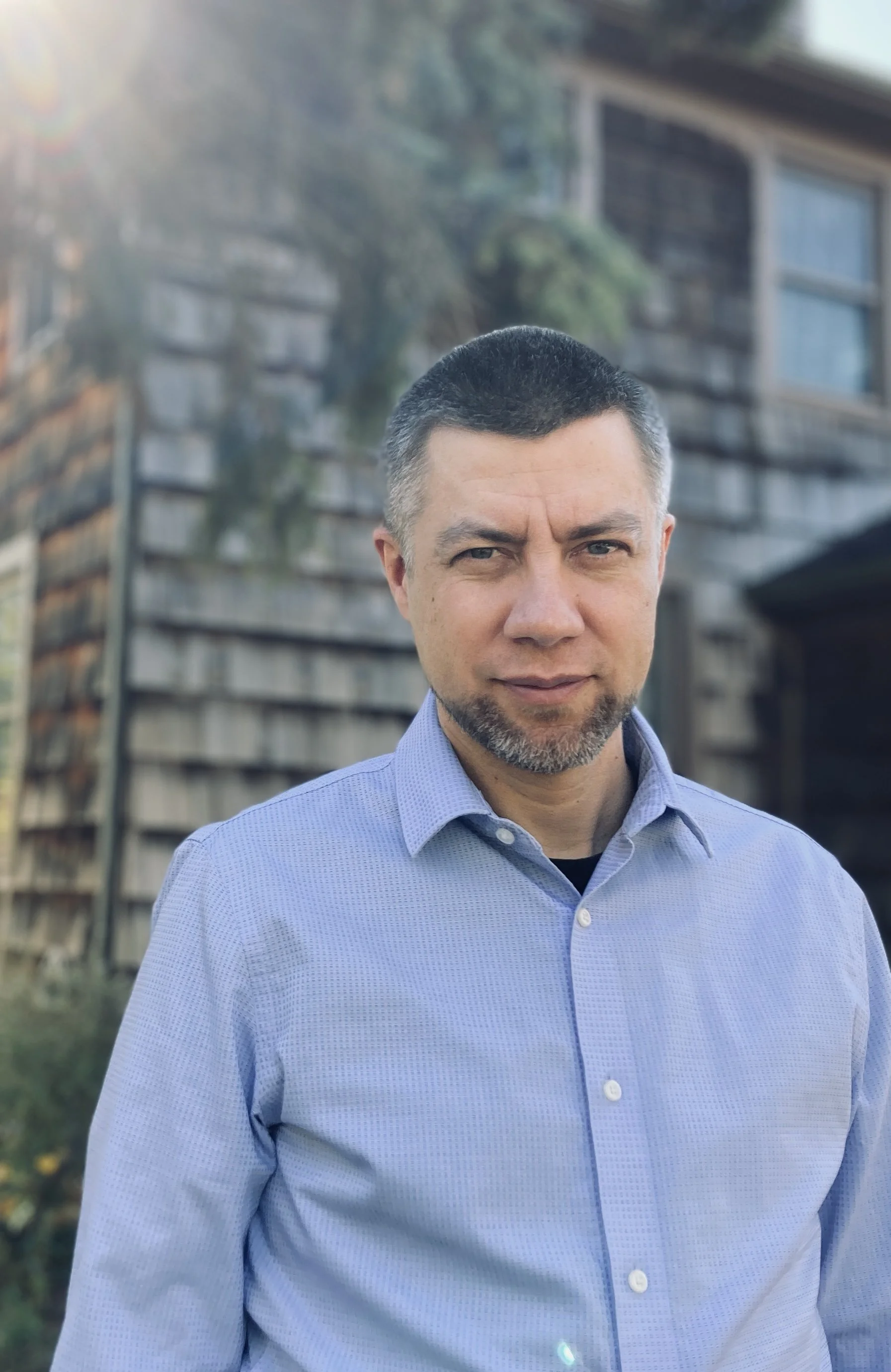 A man with short dark hair, a beard, and blue eyes wearing a light blue collared shirt stands outdoors in front of a wooden fence and trees, with sunlight shining from the left.