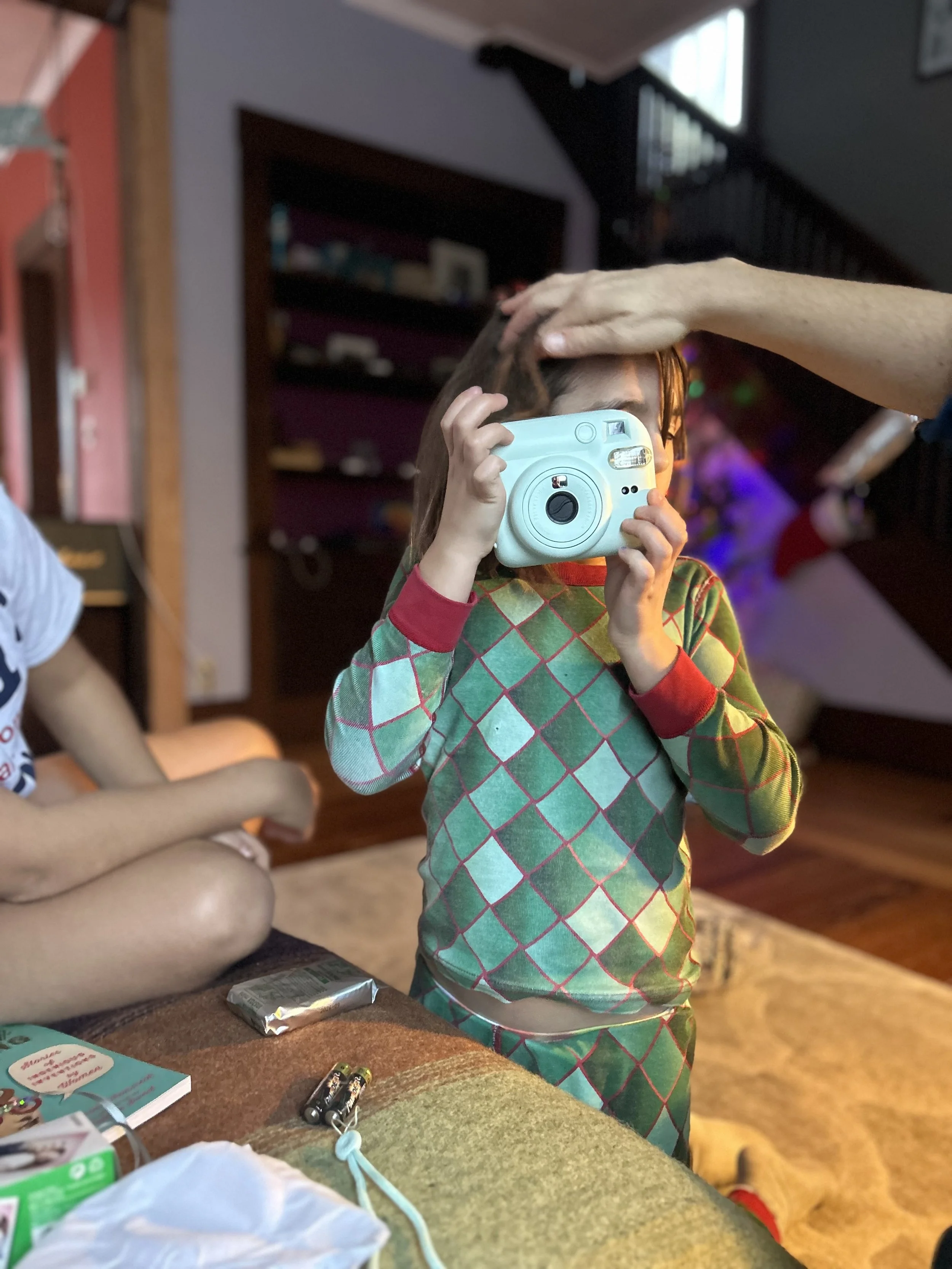A young girl wearing a colorful diamond-patterned shirt taking a photo with a disposable camera, with an adult gently placing a hand on her head, in a cozy living room with shelves and Christmas decorations in the background.