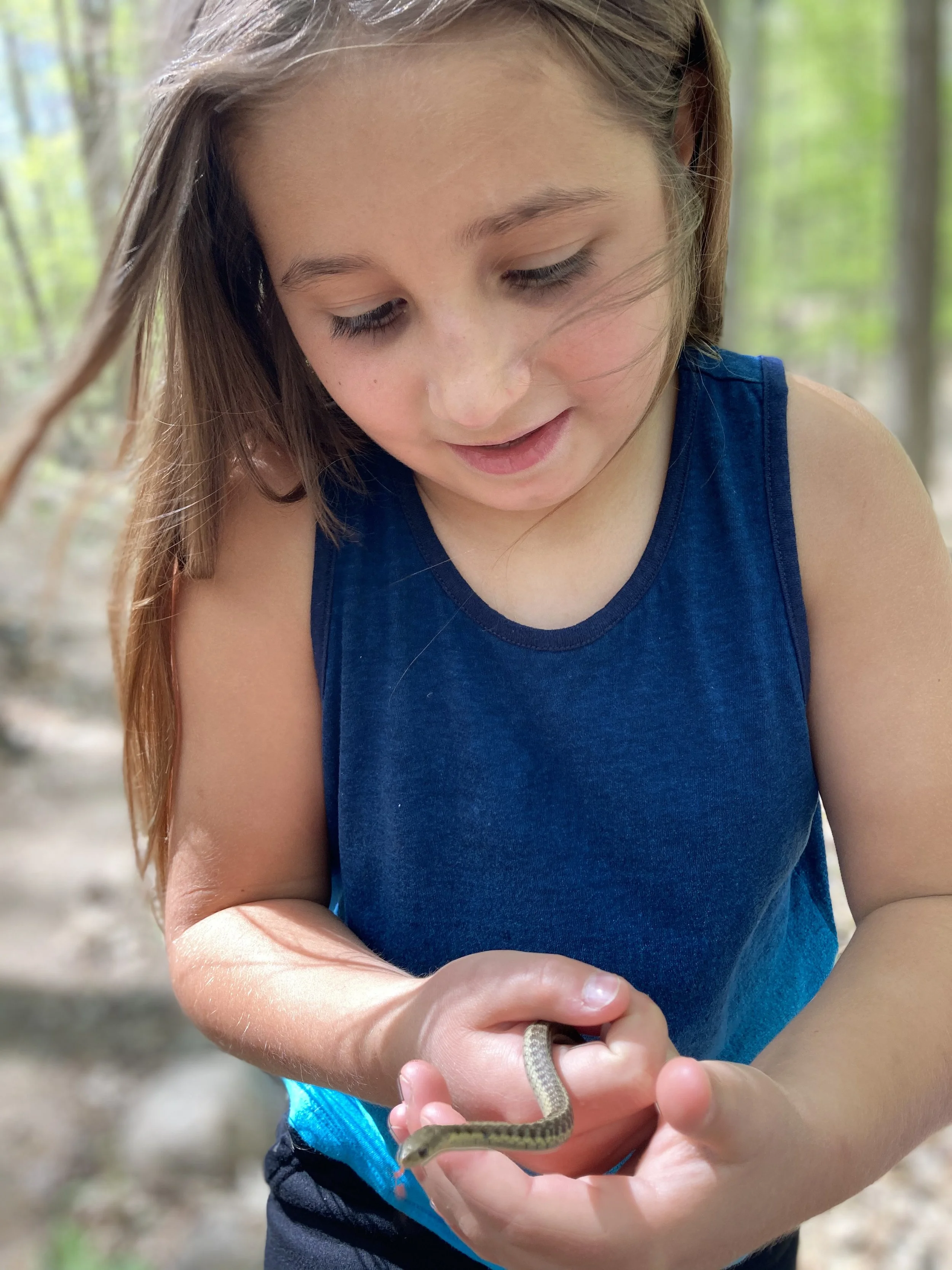 A young girl with long hair, wearing a blue sleeveless shirt, holding a small snake in her hands. The background shows a blurred outdoor wooded area.