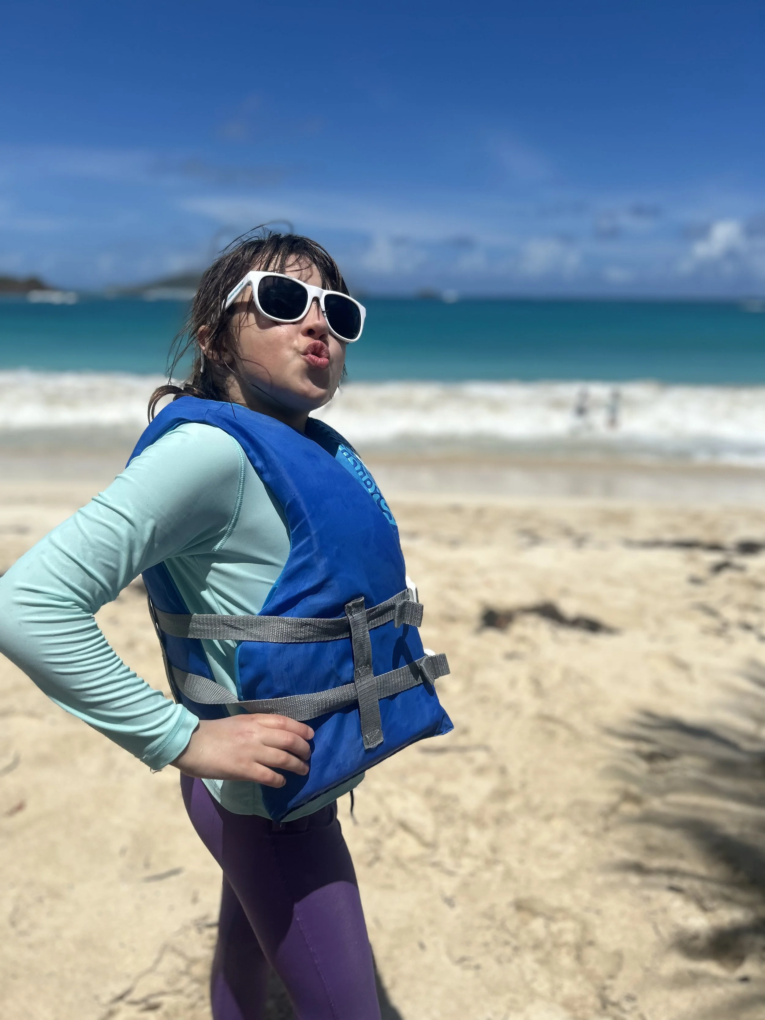 A young woman with dark hair wearing sunglasses, a teal long sleeve shirt, purple leggings, and a blue life vest standing on a sandy beach with the ocean and a partly cloudy sky in the background.